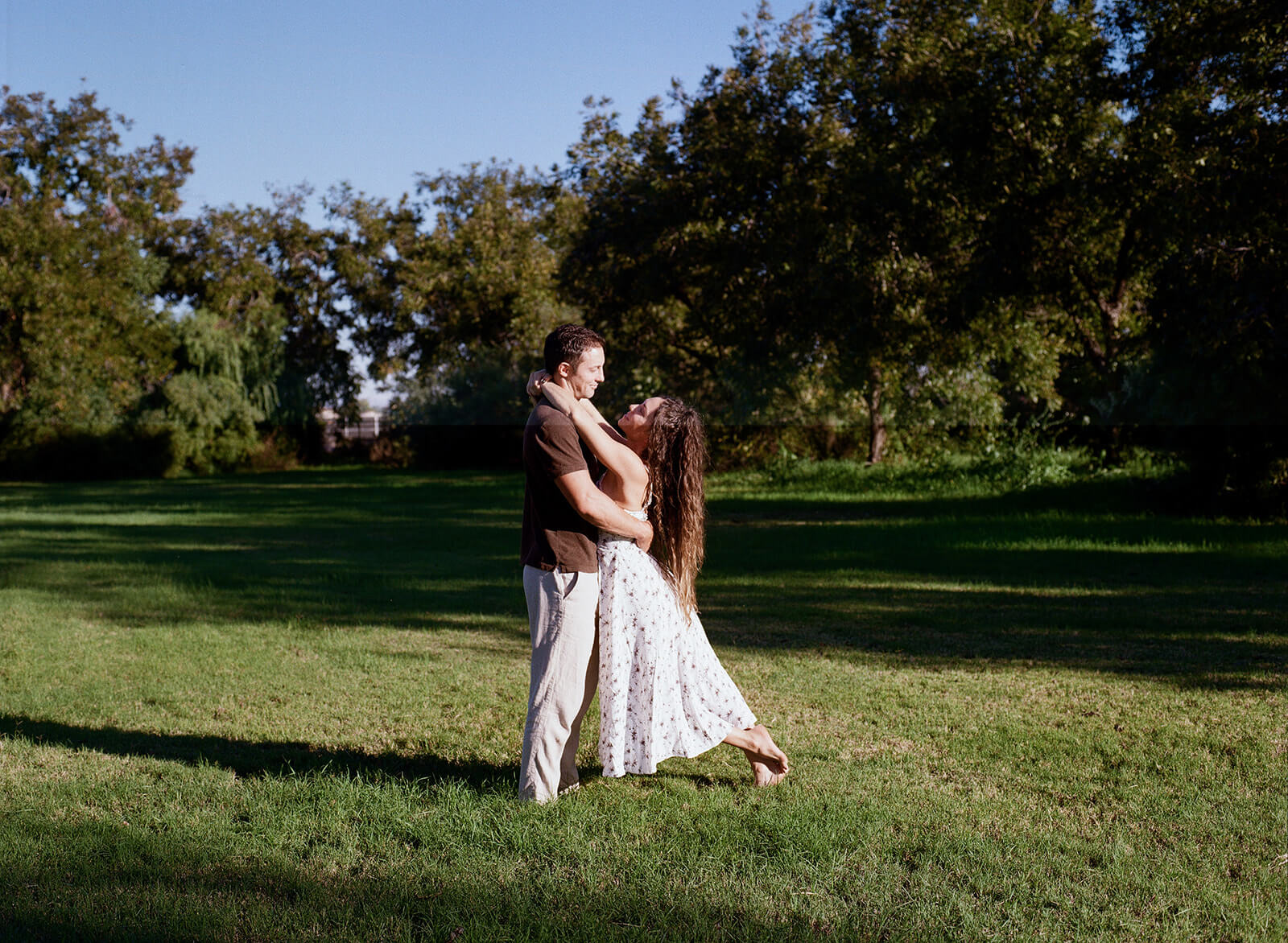 A couple dances together in an open grassy meadow at golden hour, the woman's floral dress swinging as she twirls at The Meadow in downtown Mesa.