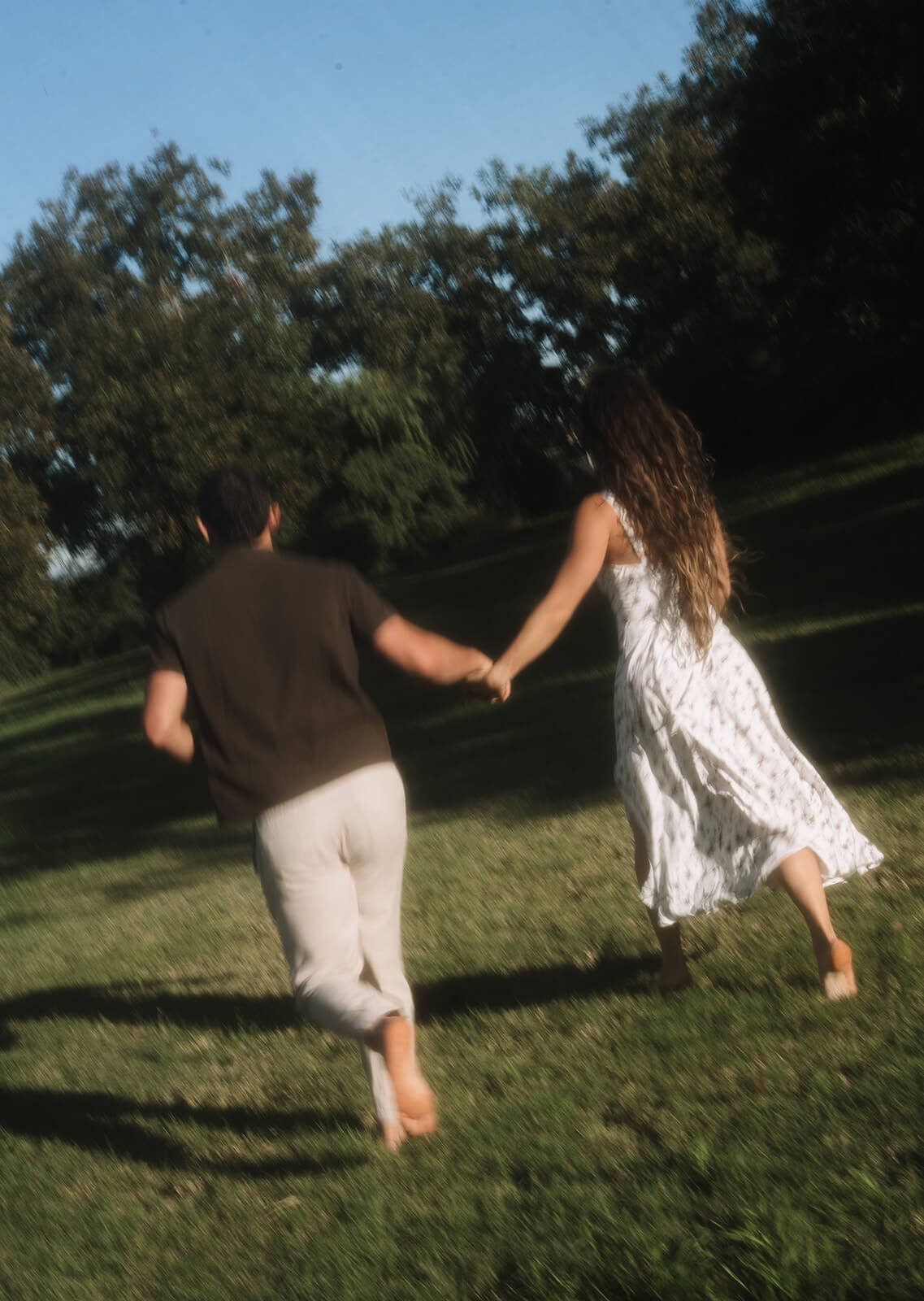 A film photo of a couple running hand in hand across a wide grassy meadow, shot from behind with motion blur and bright afternoon light, at one of Arizona's most lush garden engagement photoshoot locations.