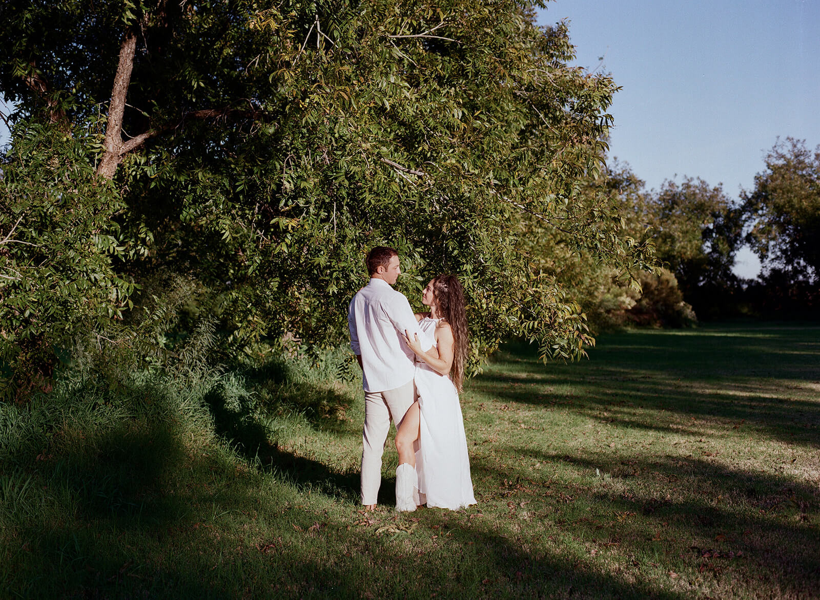 A couple stands close together beneath a large shade tree at The Meadow in Mesa Arizona, lush green grass and blue sky surrounding them at one of Arizona's most unique garden engagement photoshoot locations.