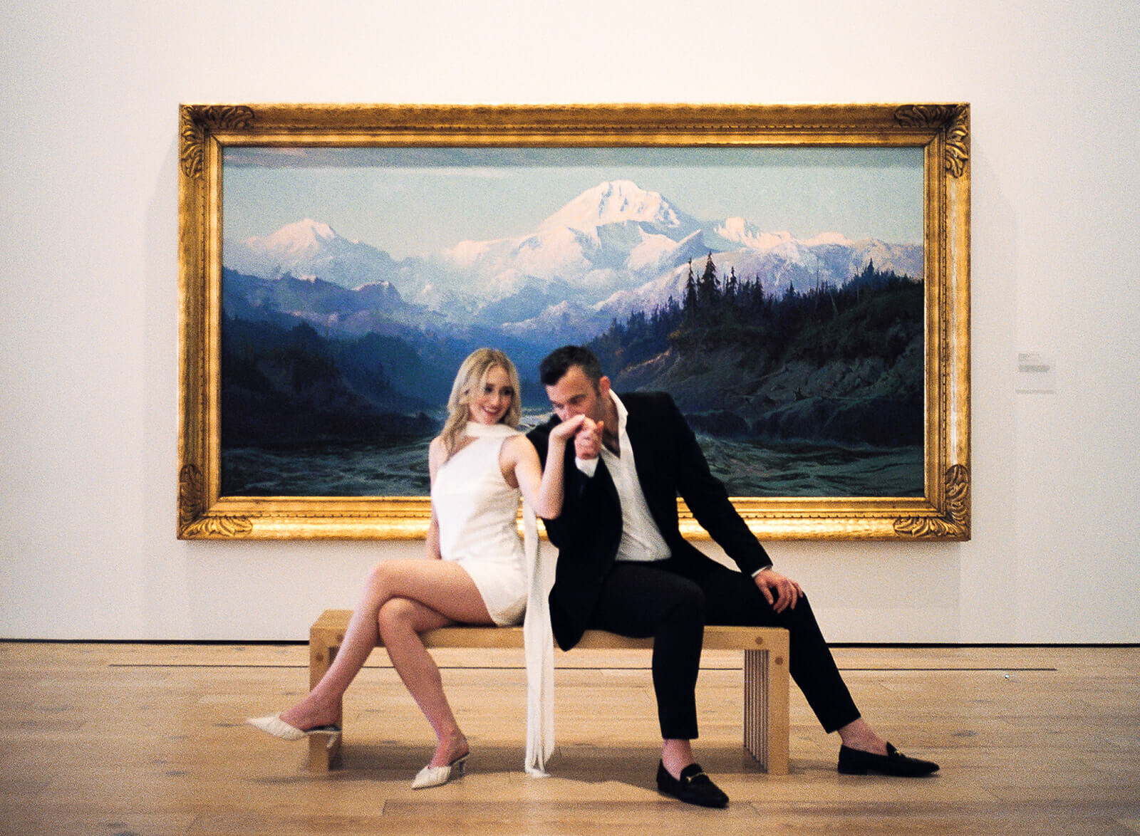 A couple sits on a gallery bench inside the Phoenix Art Museum, the man kissing the woman's hand while she smiles at him, a large gold-framed mountain landscape painting spanning the wall behind them at one of the most editorial engagement photoshoot locations in Phoenix.