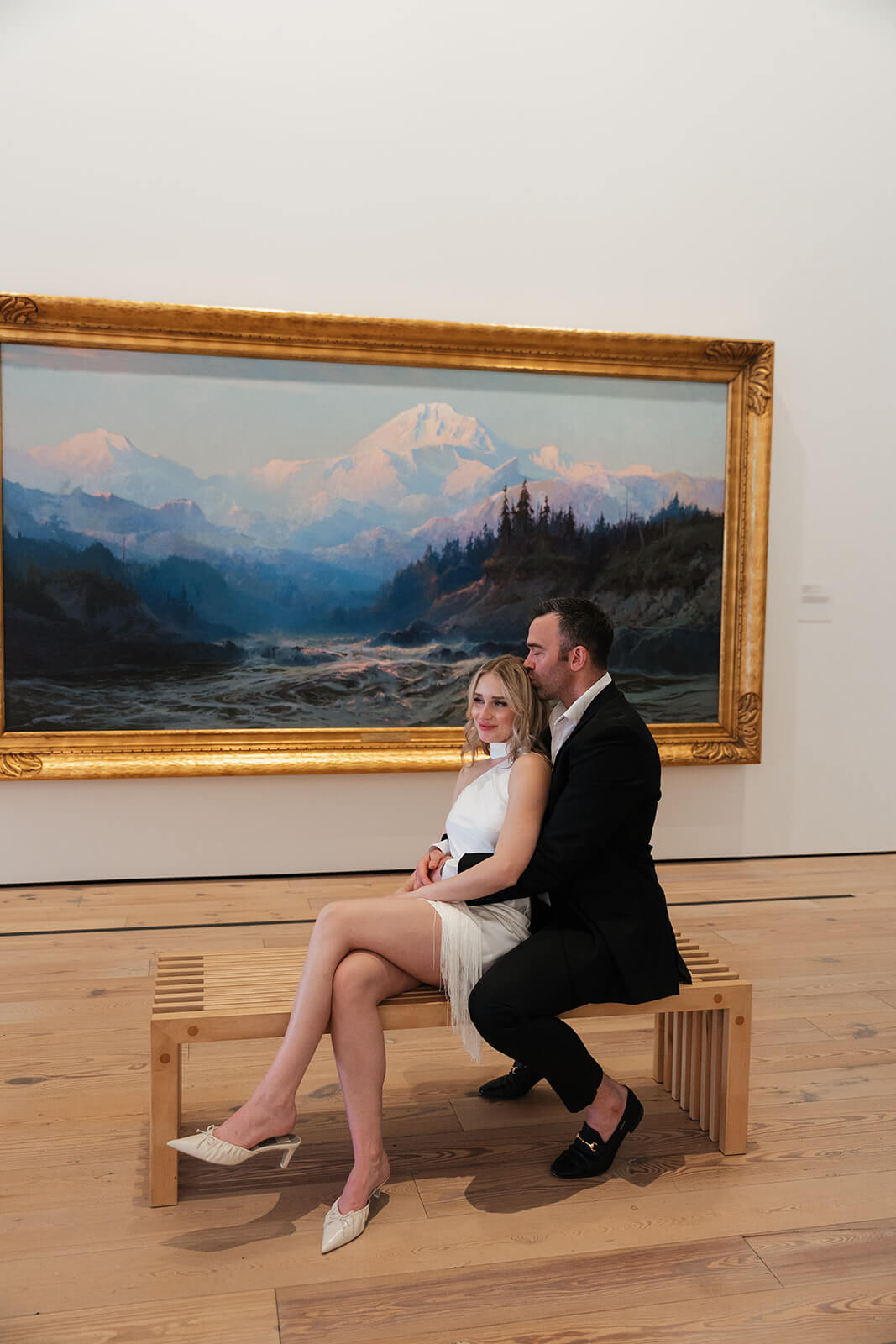A couple sits together on a wooden gallery bench inside the Phoenix Art Museum, the man whispering to the woman as she looks toward the camera, a large gold-framed mountain landscape painting filling the wall behind them at one of the most editorial engagement photoshoot locations in Arizona.