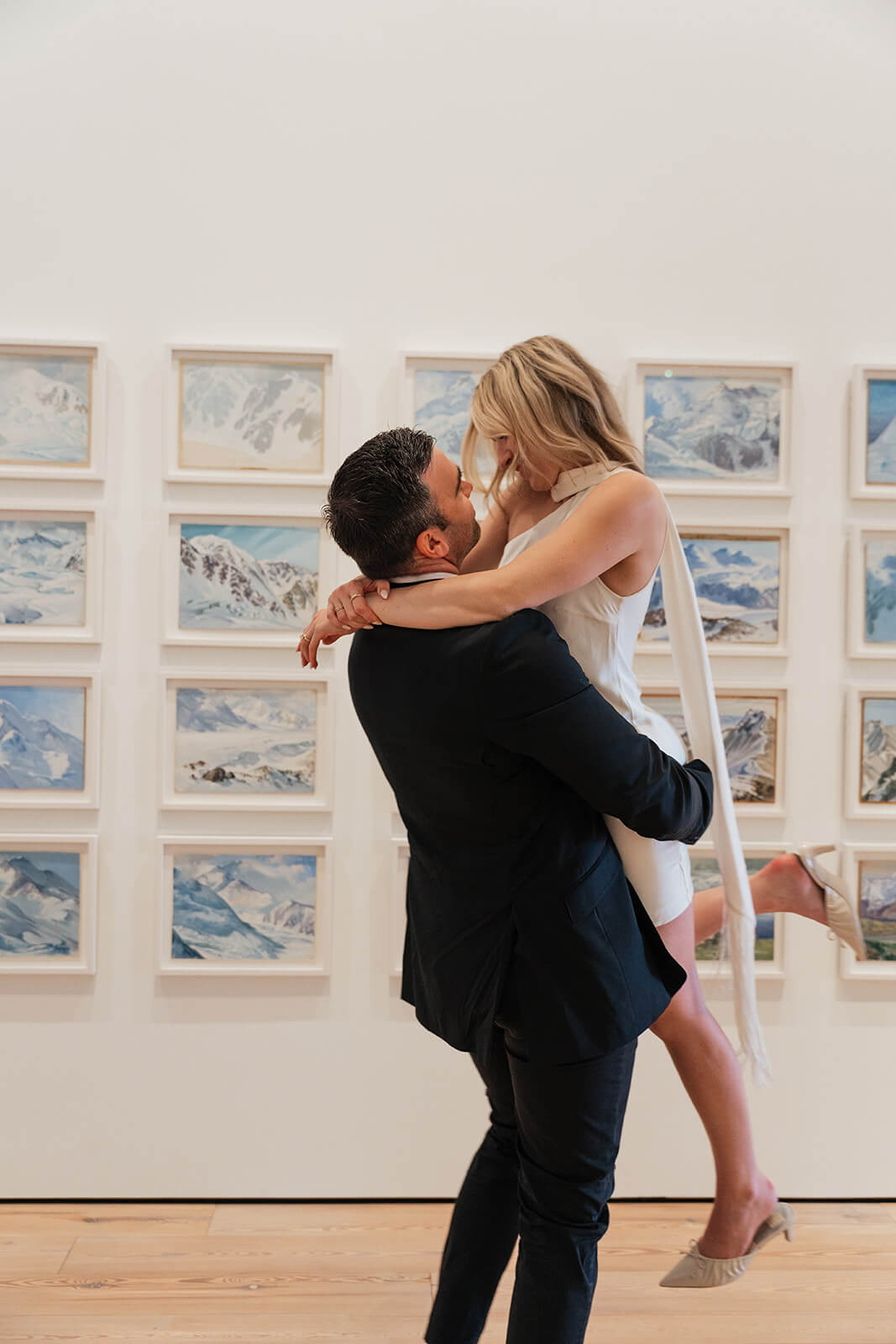 A man lifts his partner off the ground as they kiss inside the Phoenix Art Museum, a wall of framed mountain watercolors covering the white gallery wall behind them.
