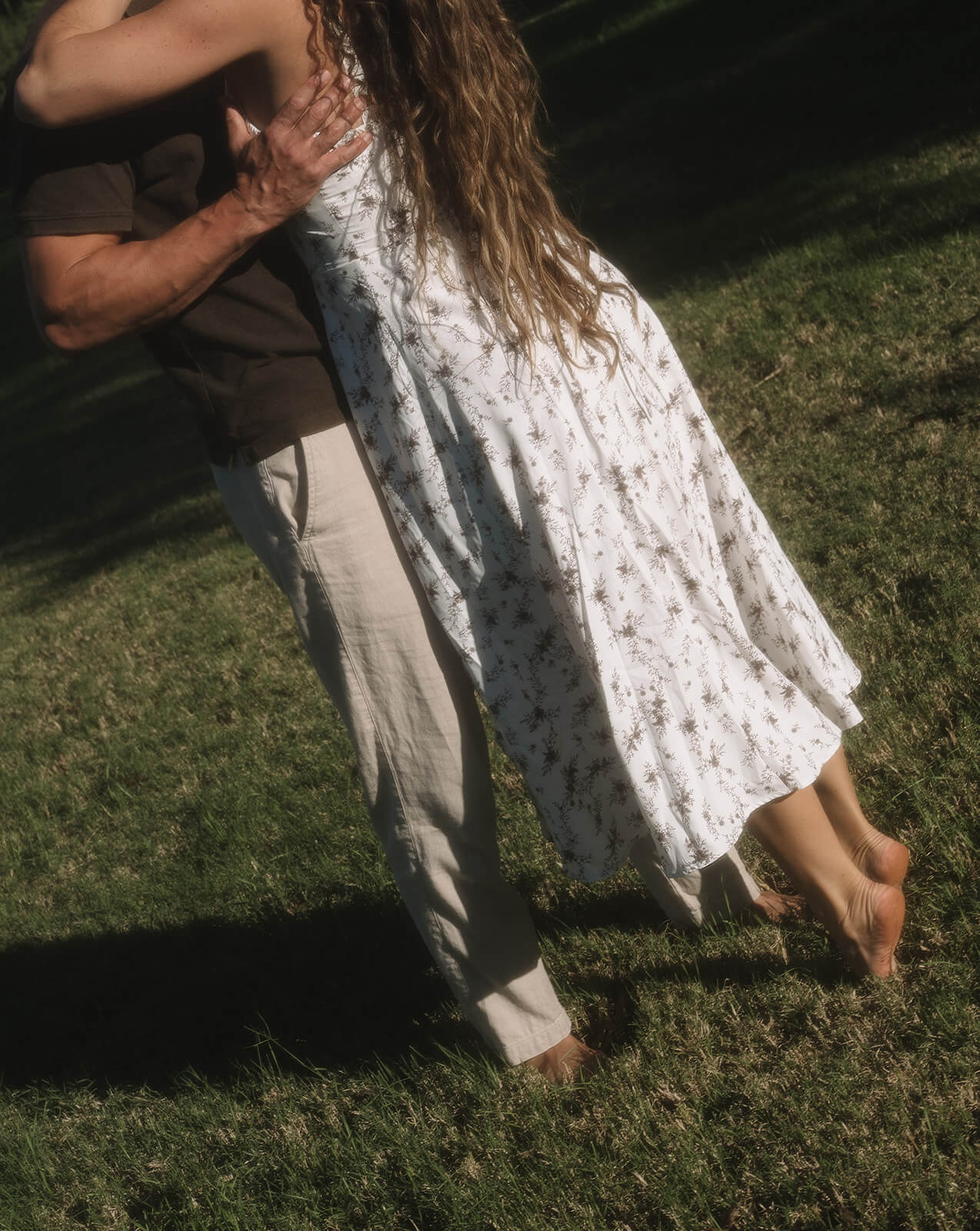 A detail shot of a couple dancing together on the grass, the woman's white floral dress swirling around her legs in warm late afternoon light at The Meadow in Mesa.