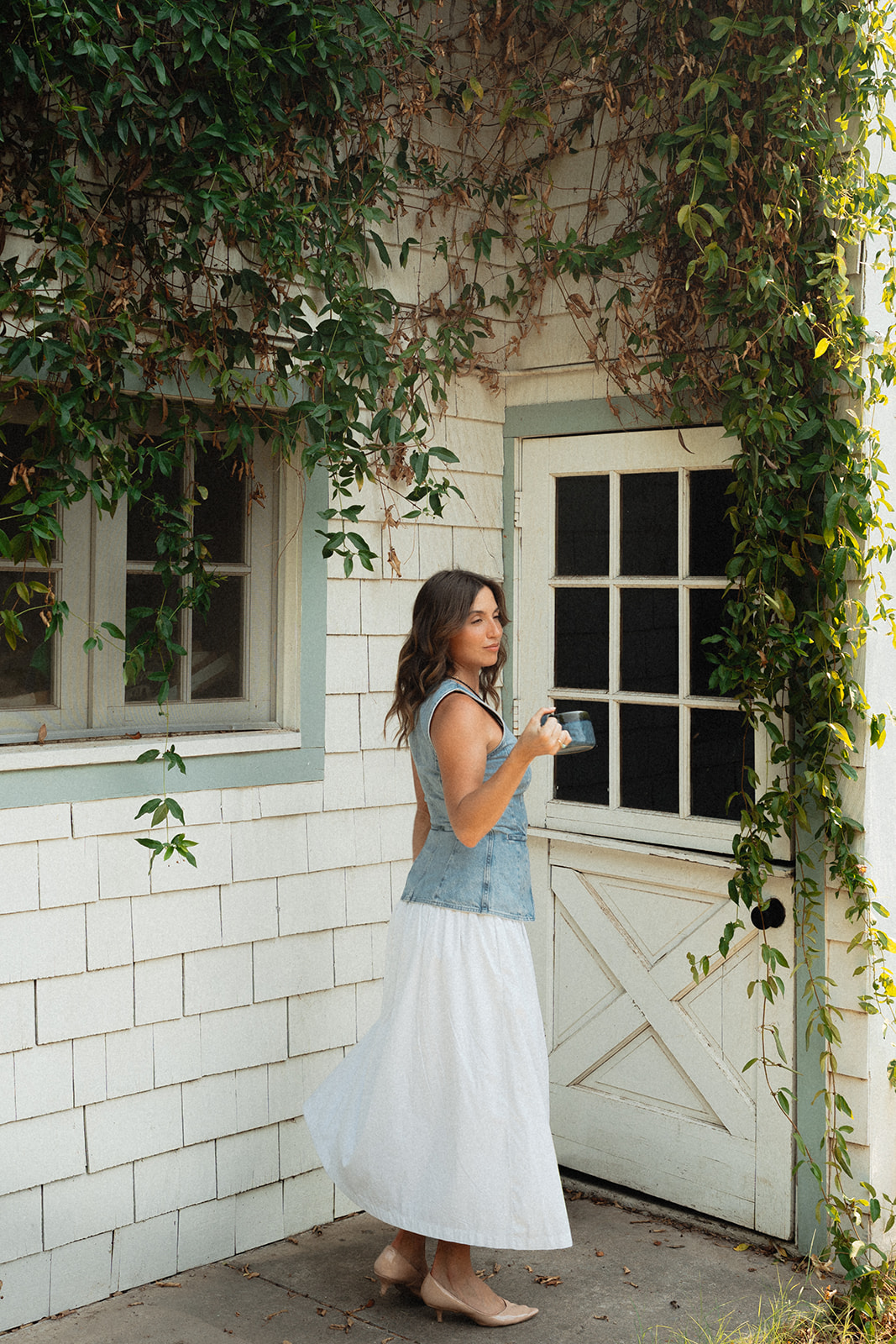 A woman in a denim vest and white skirt stands beside an ivy covered cottage door at The Bloomstead, holding a coffee cup and looking to the side.