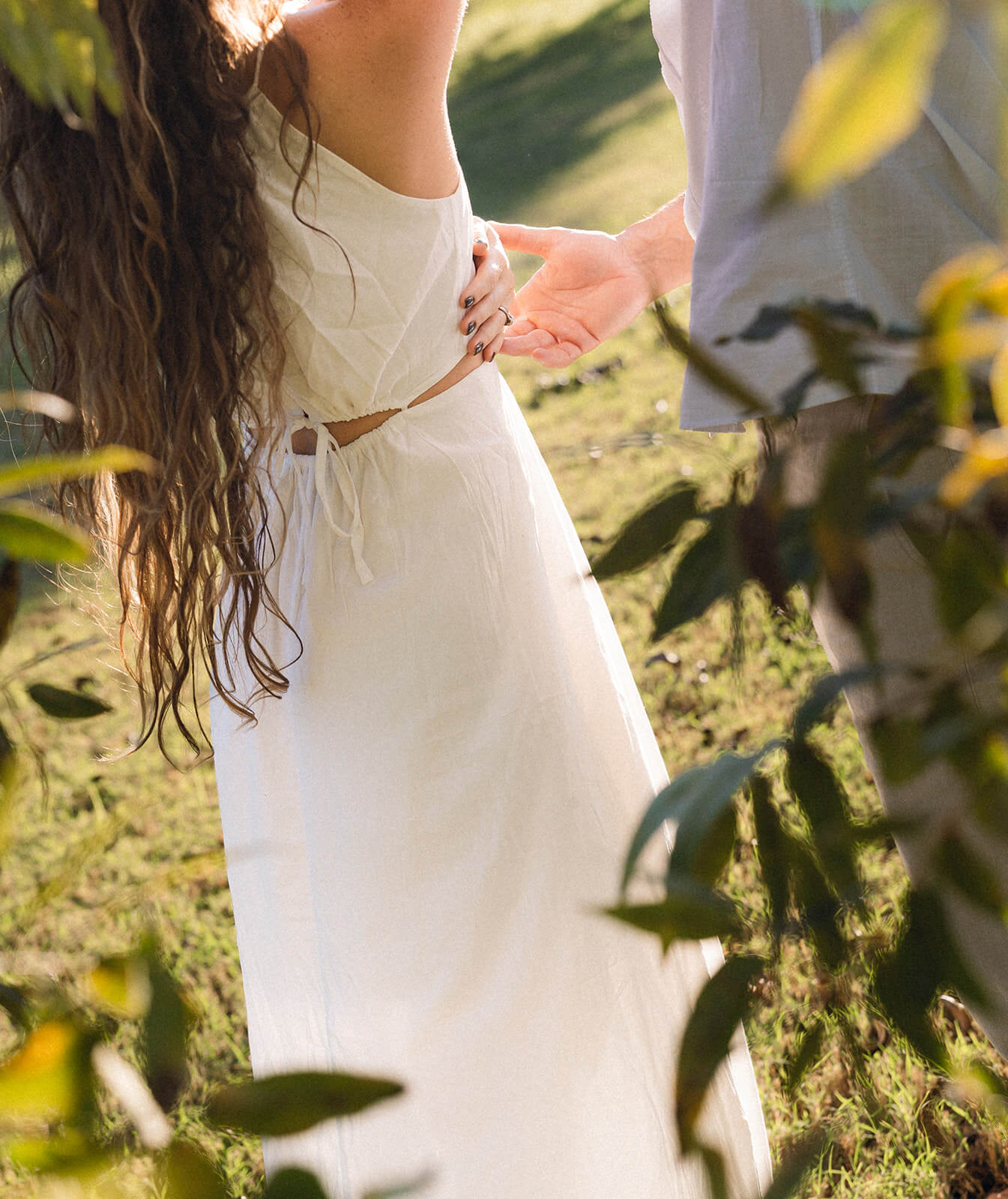 A close up detail shot of a couple holding hands at a garden engagement session in Arizona, the woman's white linen dress and long wavy hair catching the light with green foliage in the foreground.