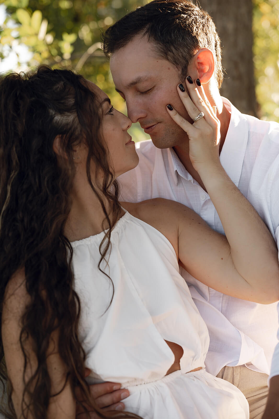 A close up of a couple almost kissing at golden hour beneath a shade tree, the woman's hand resting softly on the man's face during their garden engagement session in Arizona.
