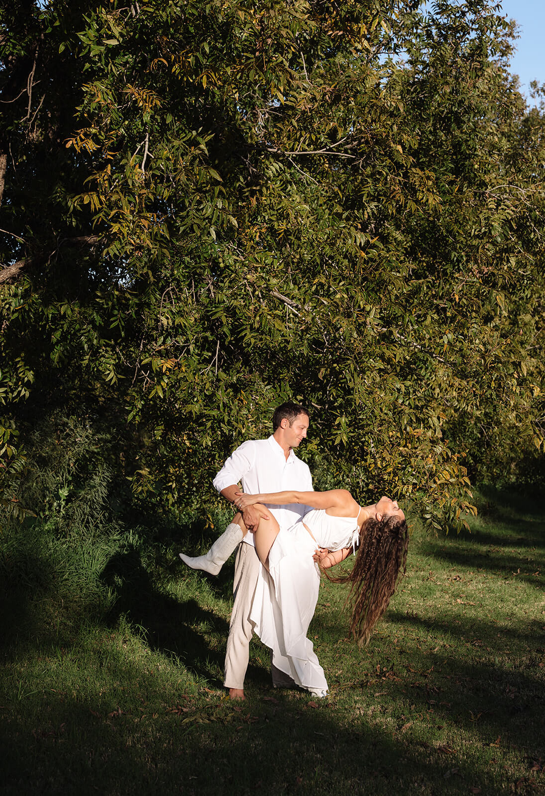 A man dips a woman low over the grass beneath a large shade tree at golden hour, both laughing at one of Arizona's most lush garden engagement photoshoot locations.