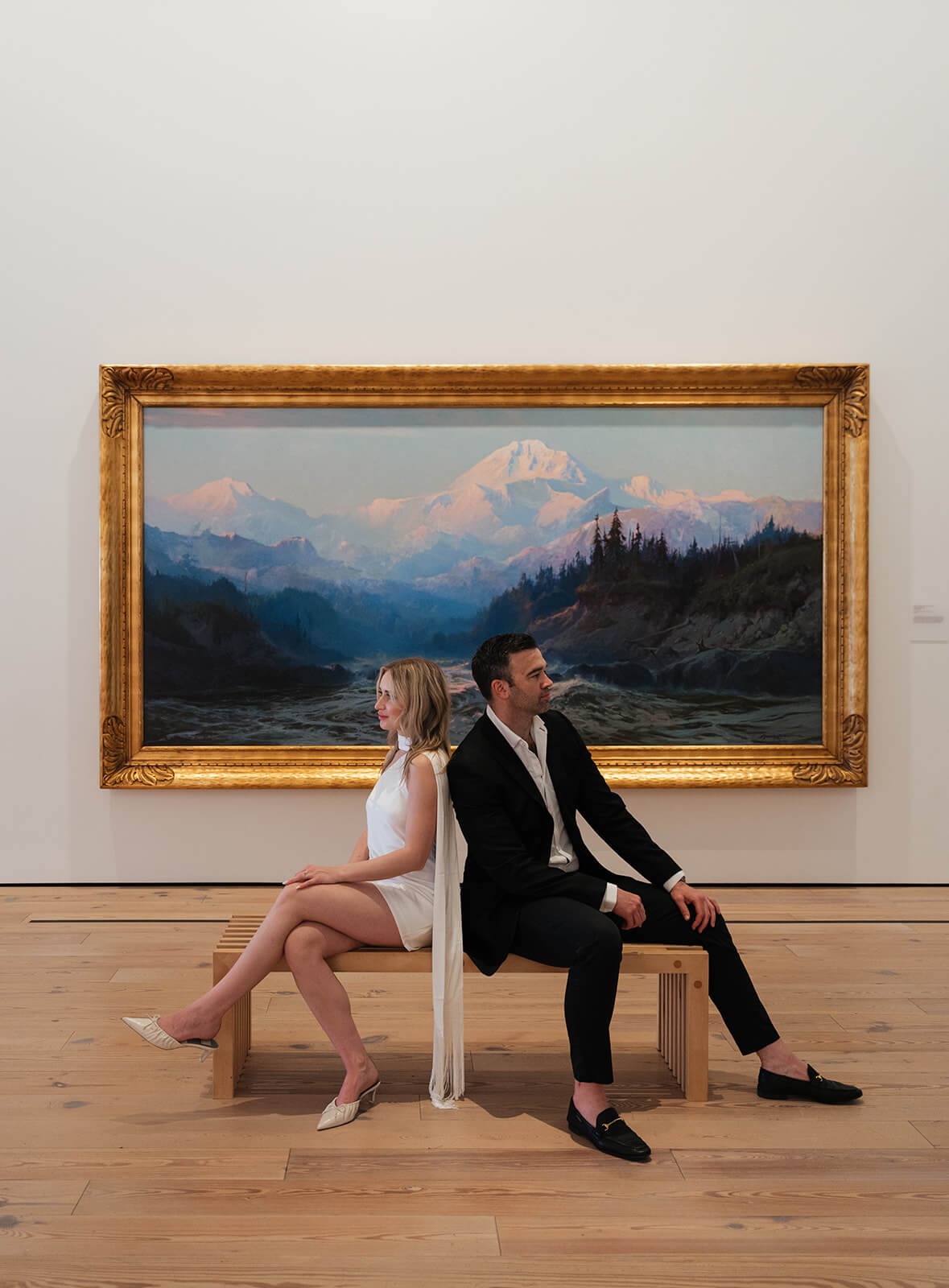 A couple sits back to back on a wooden gallery bench inside the Phoenix Art Museum, one of the most editorial engagement photoshoot locations in Arizona, a large gold-framed landscape painting filling the wall behind them.
