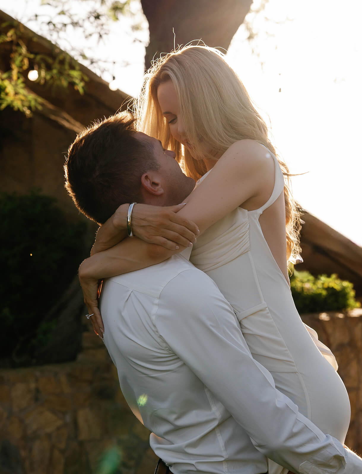 A couple kisses in warm golden backlight outdoors at Wrigley Mansion, the woman's hair catching the light as she wraps her arms around her partner's neck.