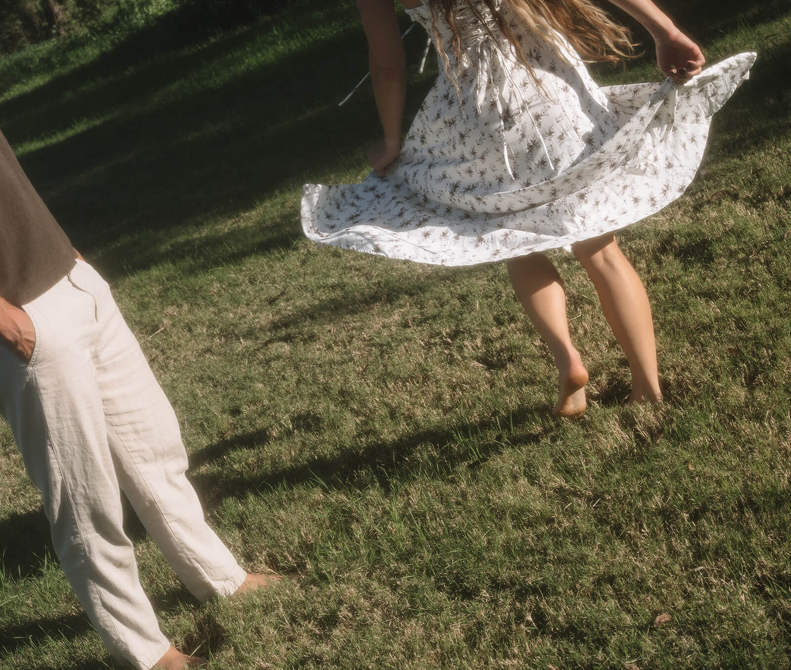 A detail shot of a couple walking barefoot across green grass at golden hour, the woman's white floral dress swirling with movement at The Meadow in Mesa, one of the most lush garden engagement photoshoot locations in Arizona.