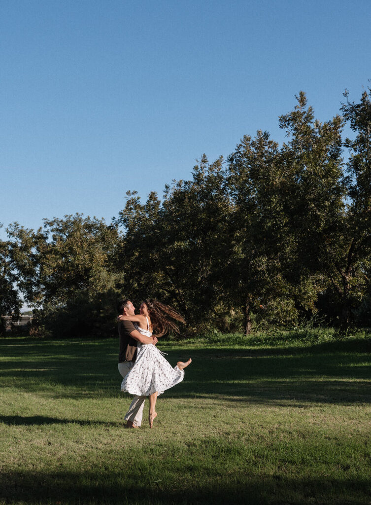 A wide shot of a man lifting and spinning a woman across a wide open green meadow under a bright blue sky, mature trees lining the edge of the field at one of Arizona's most scenic engagement photoshoot locations.