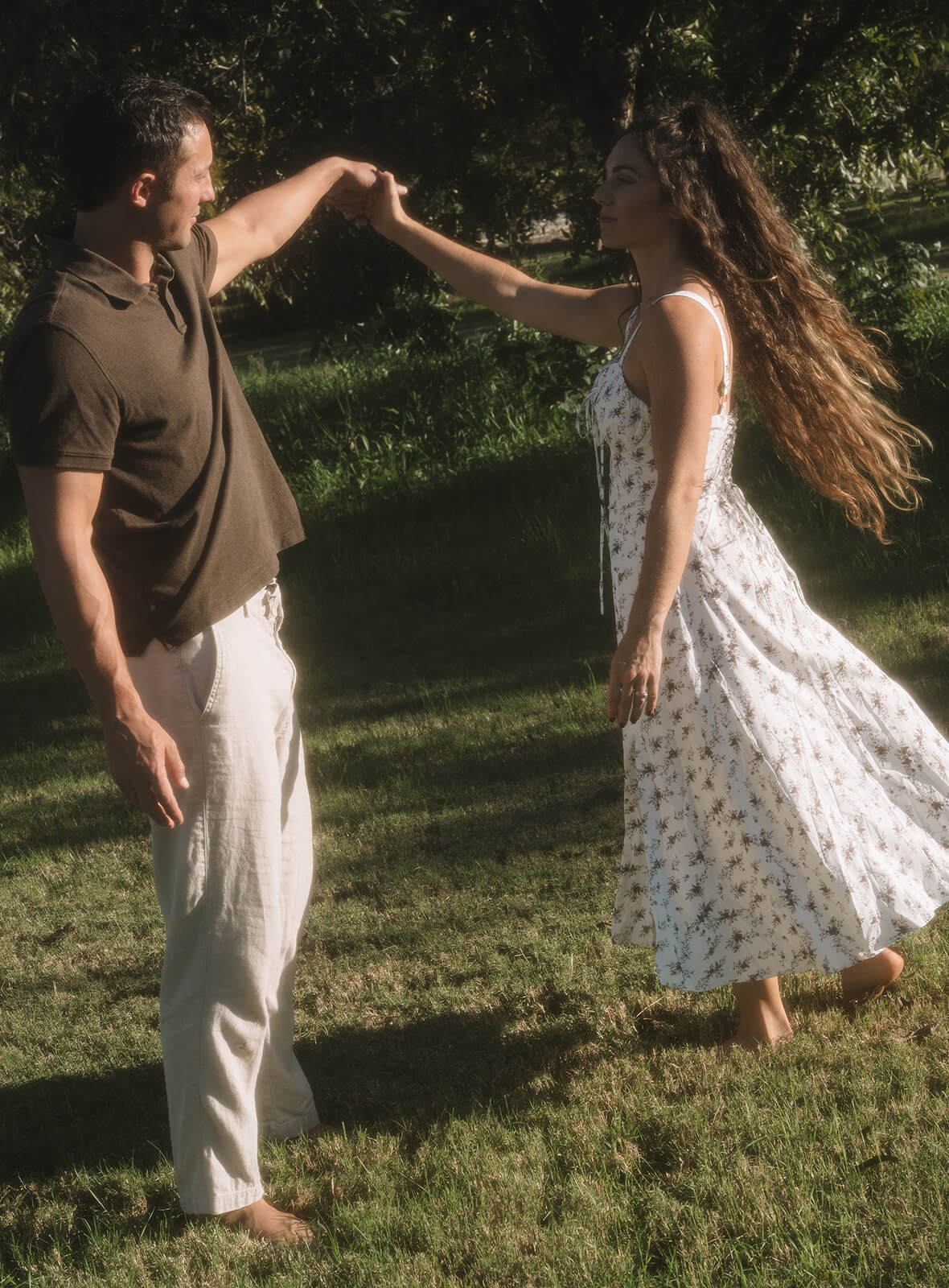 A man spins a woman barefoot on a grassy meadow at golden hour, her long wavy hair and white floral dress sweeping through the air at The Meadow in downtown Mesa.