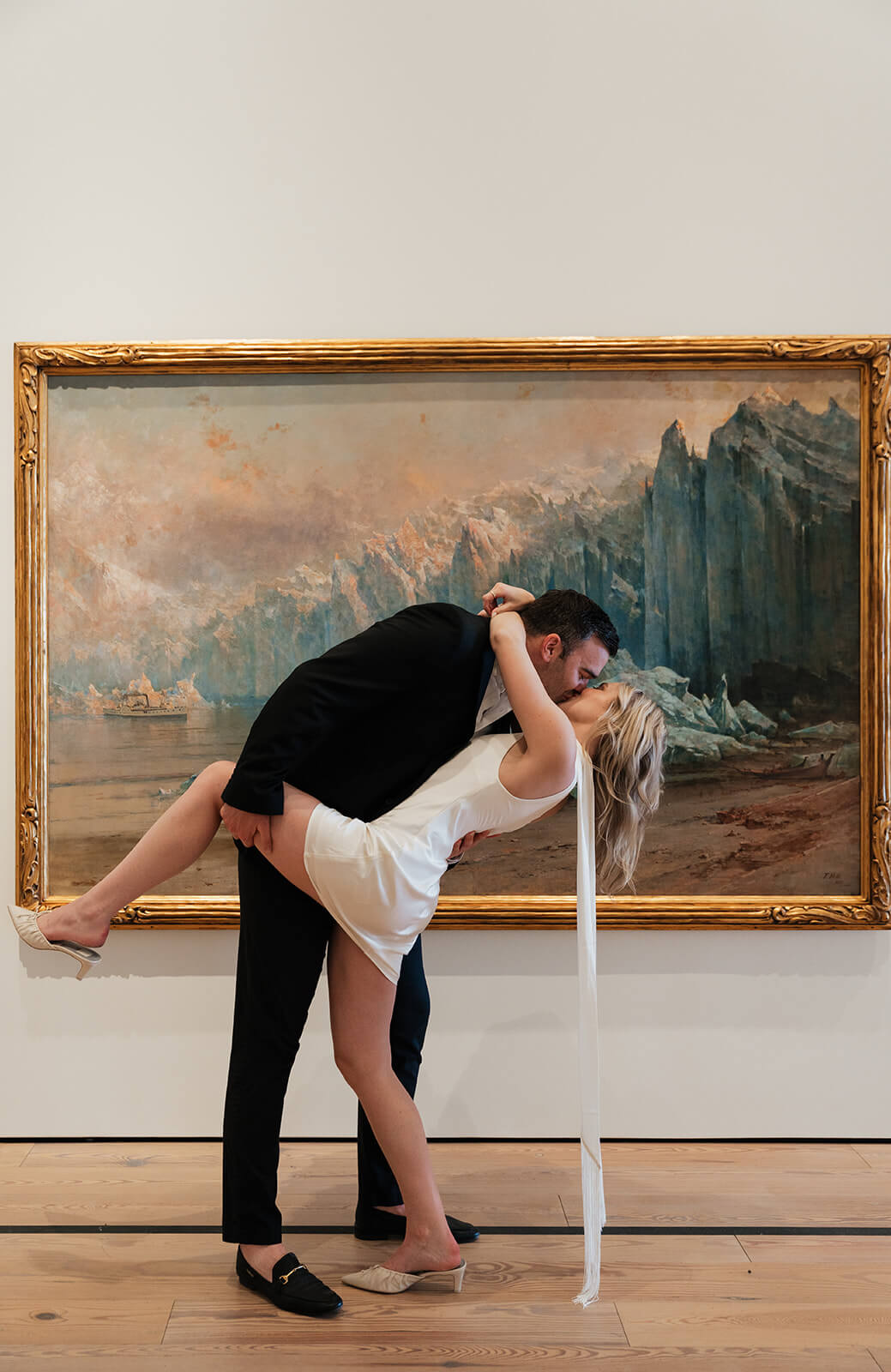 A man dips a woman dramatically in front of a large gold-framed landscape painting inside the Phoenix Art Museum, her white fringe dress catching the light on the gallery floor.