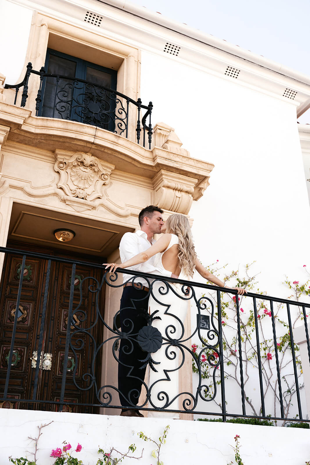 A couple kisses on an ornate wrought iron balcony on the exterior of Wrigley Mansion, the carved stone facade and pink bougainvillea framing the shot from below.