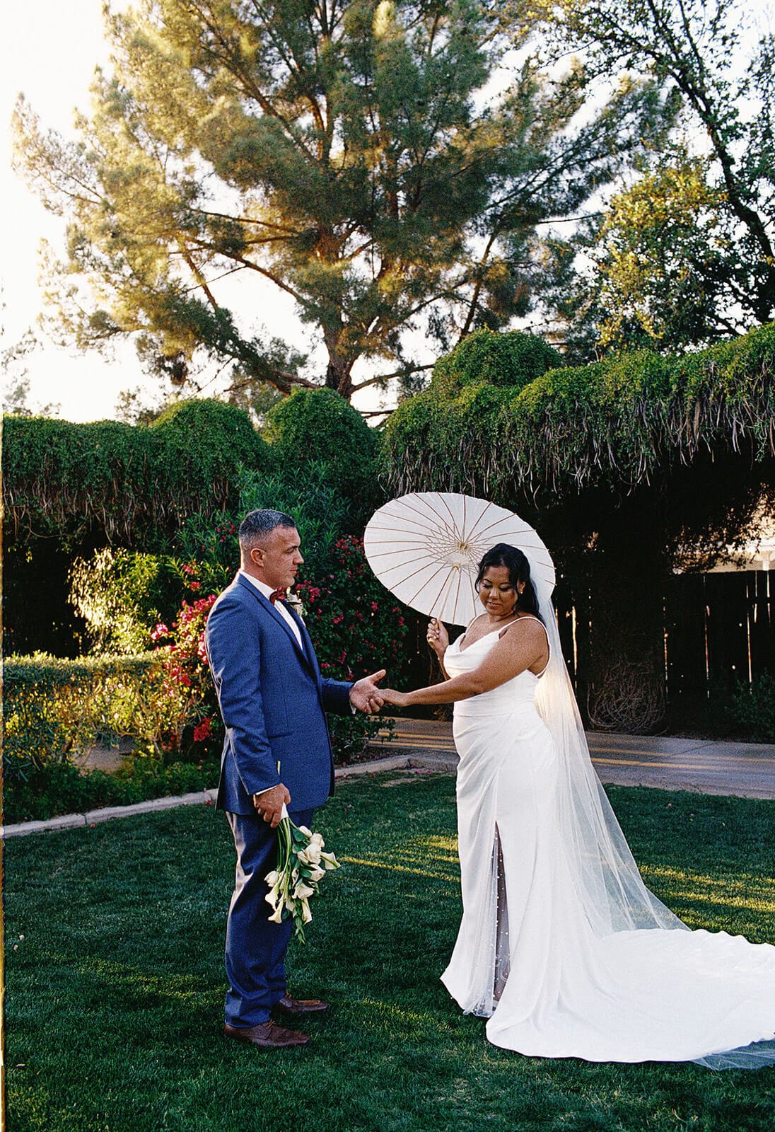 Bride holding a parasol walking with her escort through the garden grounds at Shenandoah Mill wedding venue in Gilbert Arizona
