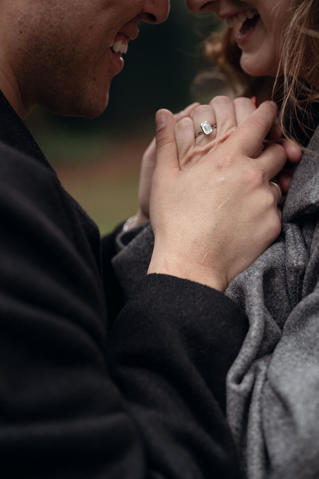 Close-up of a couple holding hands, a large diamond engagement ring prominently displayed on the woman's finger, their smiling faces softly blurred in the background.
