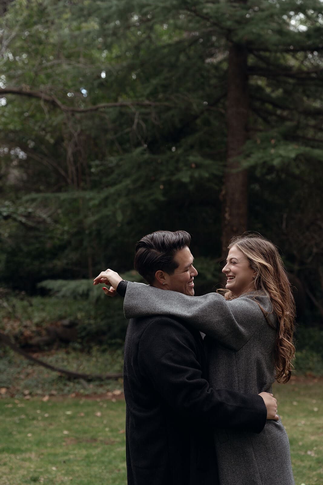 A newly engaged couple embraces and laughs together on a green lawn in front of a wooded backdrop.

