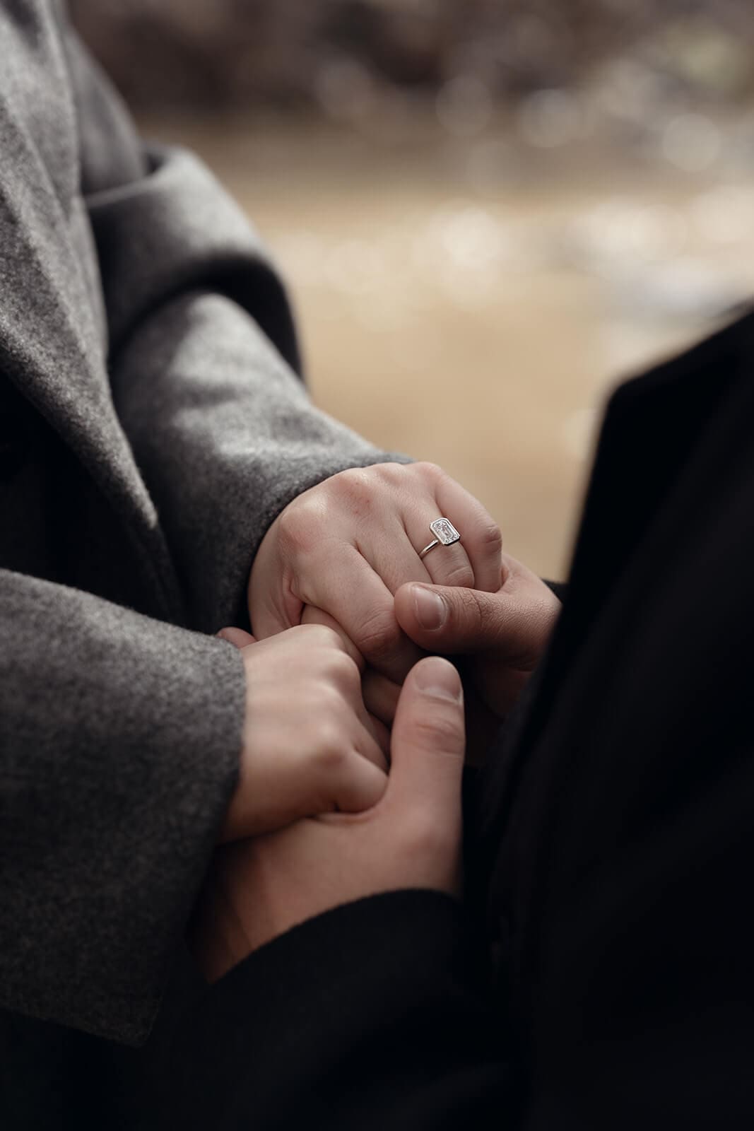 Close-up of two people holding hands, with a diamond solitaire engagement ring visible on the woman's finger, set against a blurred natural background.
