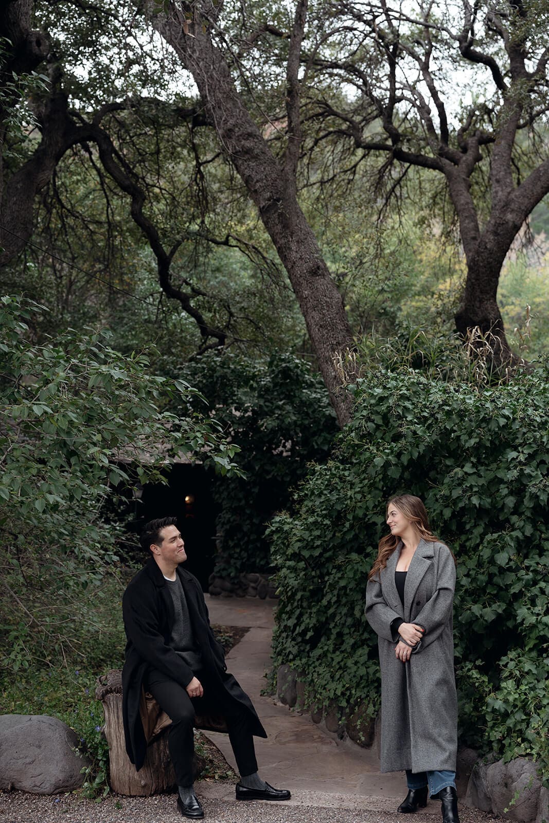 A couple sits apart on a garden path lined with lush greenery and twisted oak trees, smiling at each other in a candid, relaxed moment.
