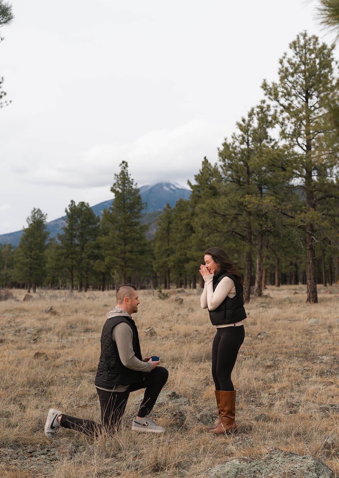A closer shot of the man on one knee holding the ring box open toward the woman, who covers her mouth in disbelief, with pine trees and mountains behind them.
