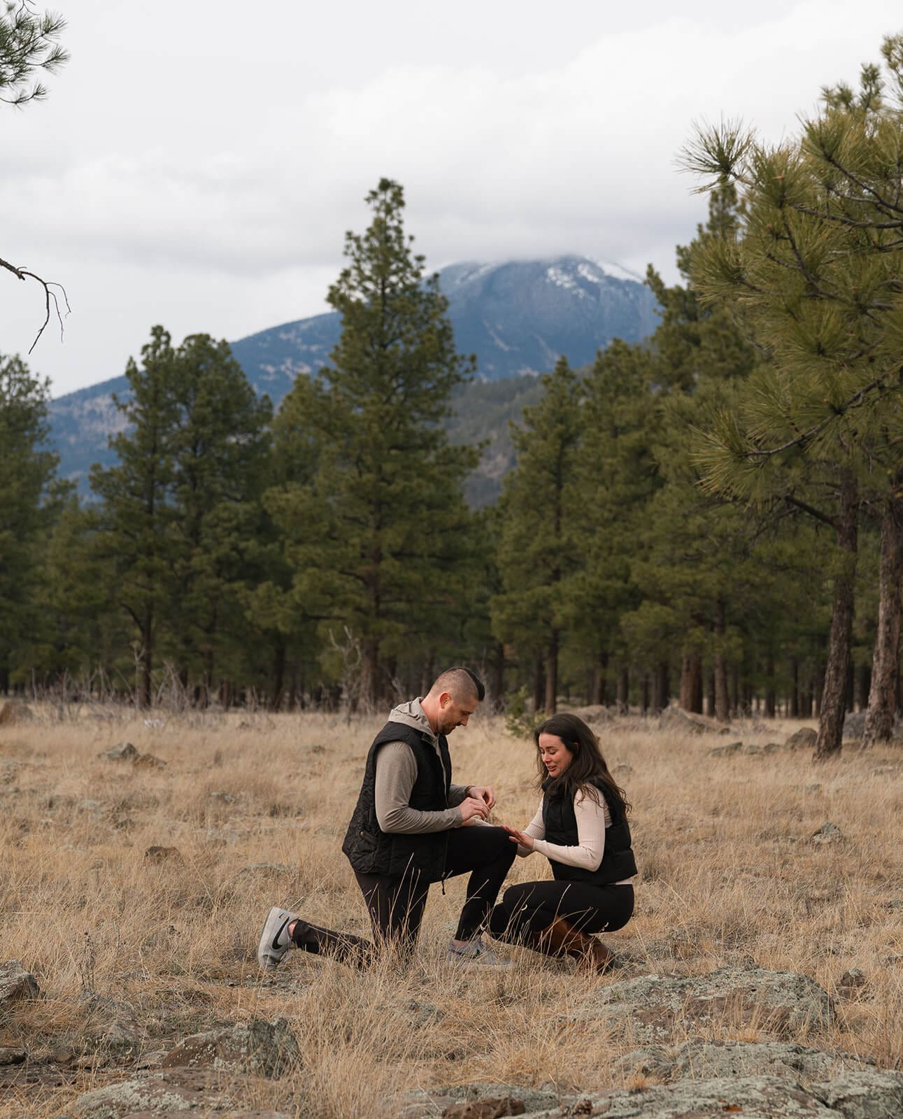 A man on one knee proposes to a woman in a dry grass meadow surrounded by pine trees and snow-capped mountains in the background.
