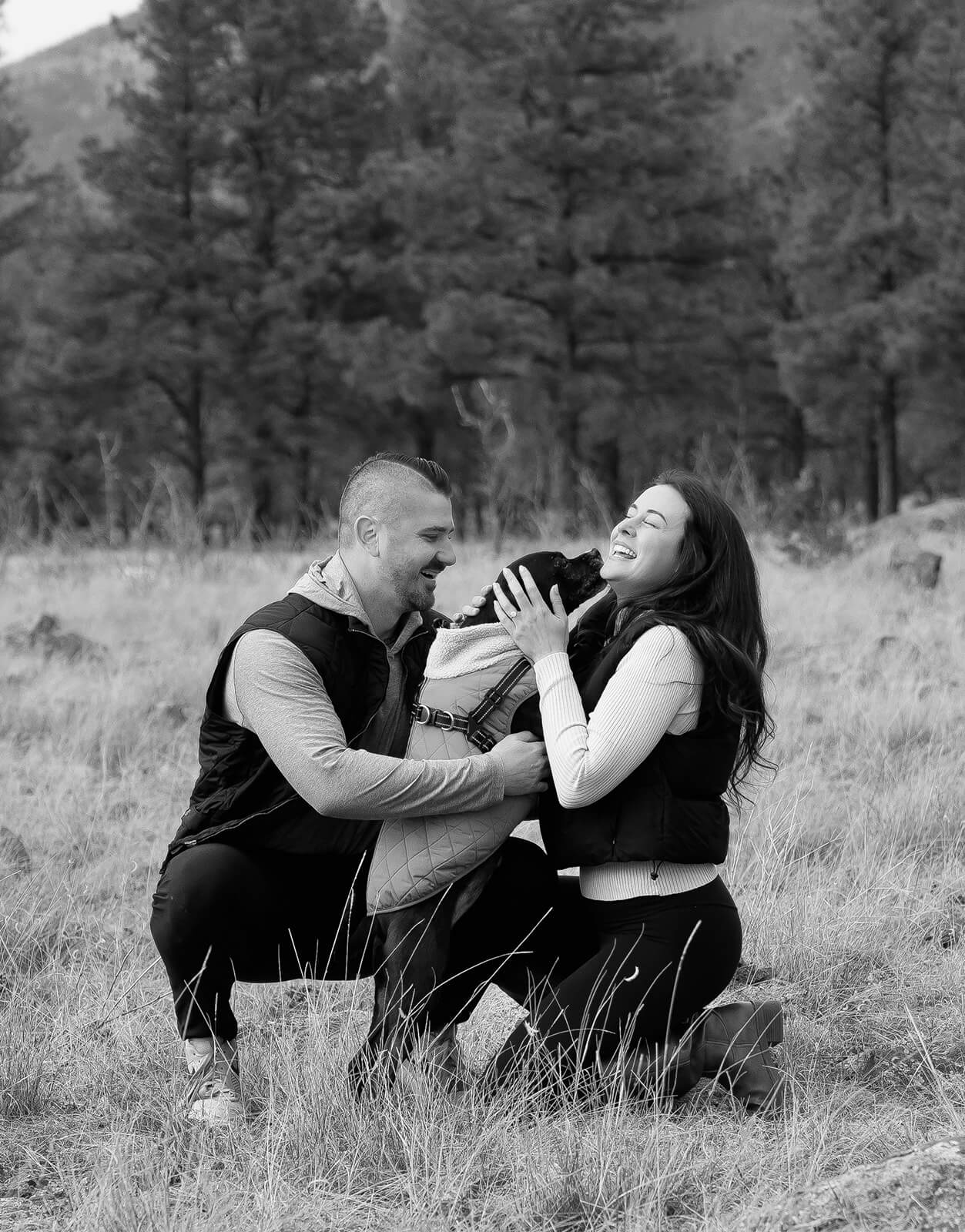 Black and white photo of a newly engaged couple kneeling together in a field, laughing joyfully while holding their dog between them.
