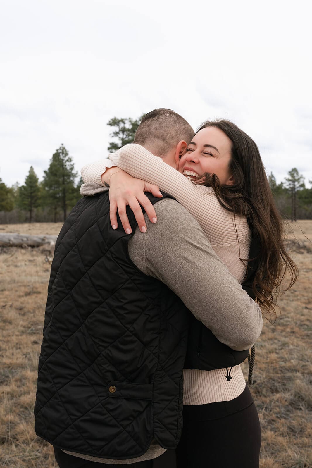 The newly engaged couple shares a tight embrace in the meadow, the woman laughing with joy as she hugs him.
