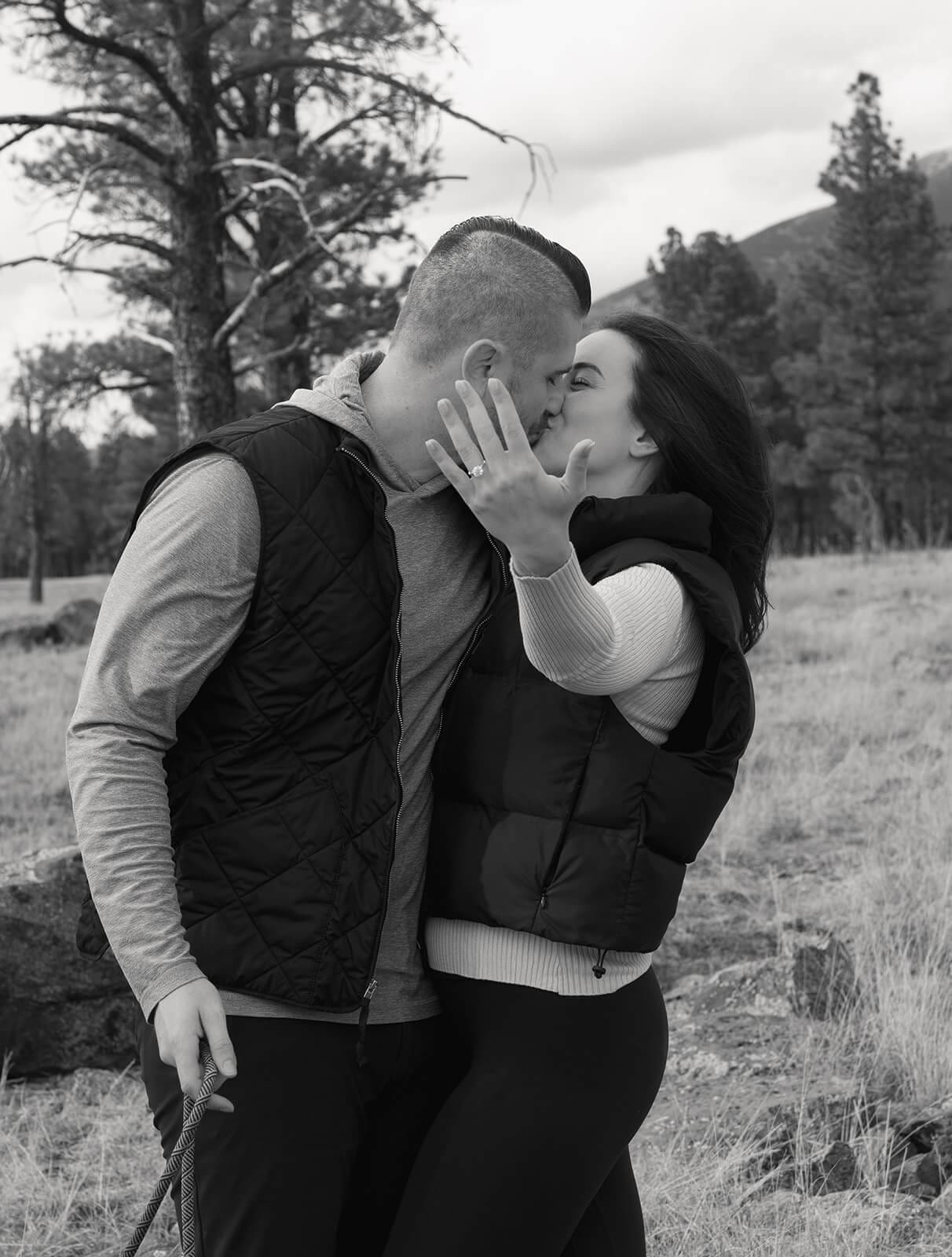 Black and white photo of the couple sharing a kiss, the woman's hand with her new engagement ring visible against his face.
