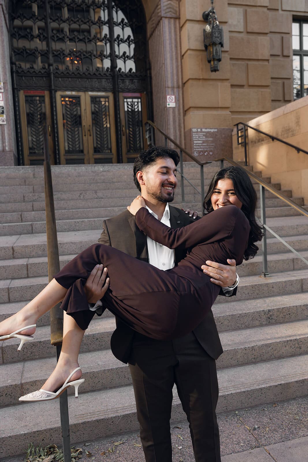 A man in a suit carries a woman in a dark dress in his arms on the steps of a stone building, both smiling joyfully.