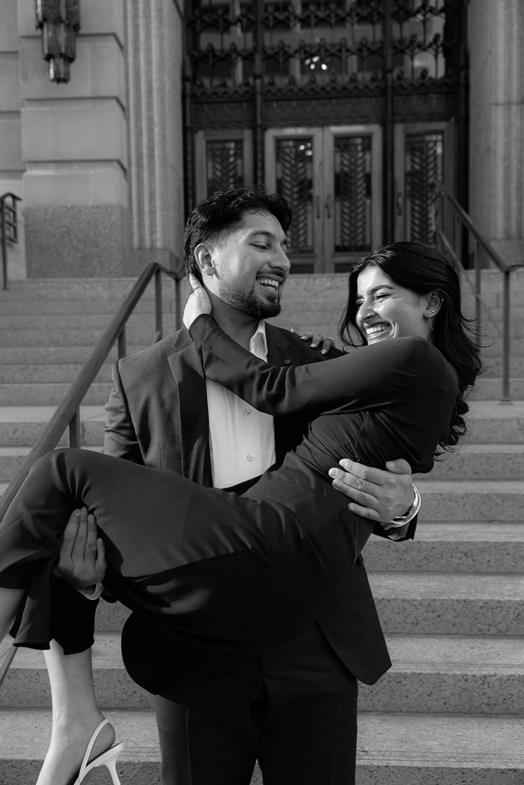 Black and white photo of a man sweeping a woman off her feet on the steps of a grand building, both laughing joyfully.
