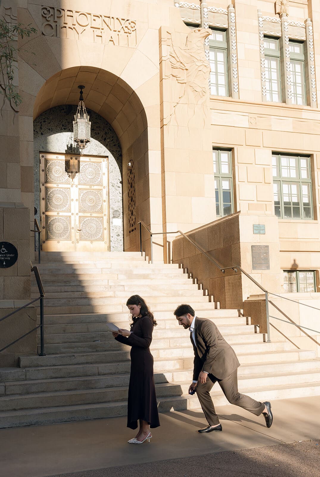 A wide shot of a man beginning to drop to one knee outside Phoenix City Hall, the woman walking ahead unaware, captured from a distance on the sunlit steps.