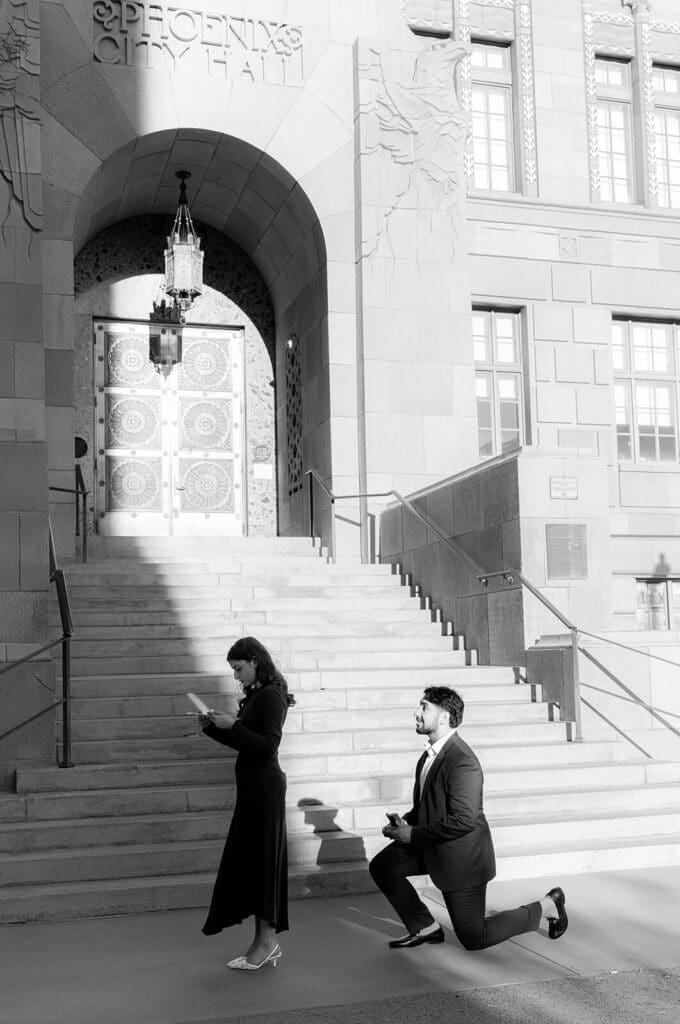 Man down on one knee proposing on the steps of a historic building in Downtown Phoenix while his partner covers her mouth in shock, with additional proposal and just-engaged portraits from Sedona's red rocks and a wooded garden setting