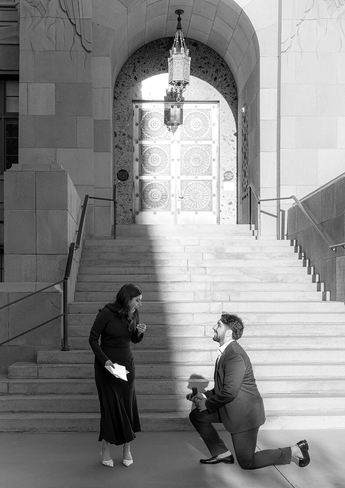 Black and white photo of a man on one knee proposing beneath the ornate arched entrance of Phoenix City Hall, the woman facing him in surprise.
