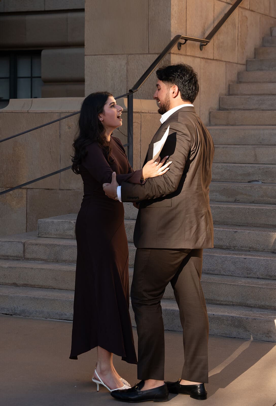 A couple stands close together on the stone steps of a grand building, gazing at each other in a tender moment after the proposal.
