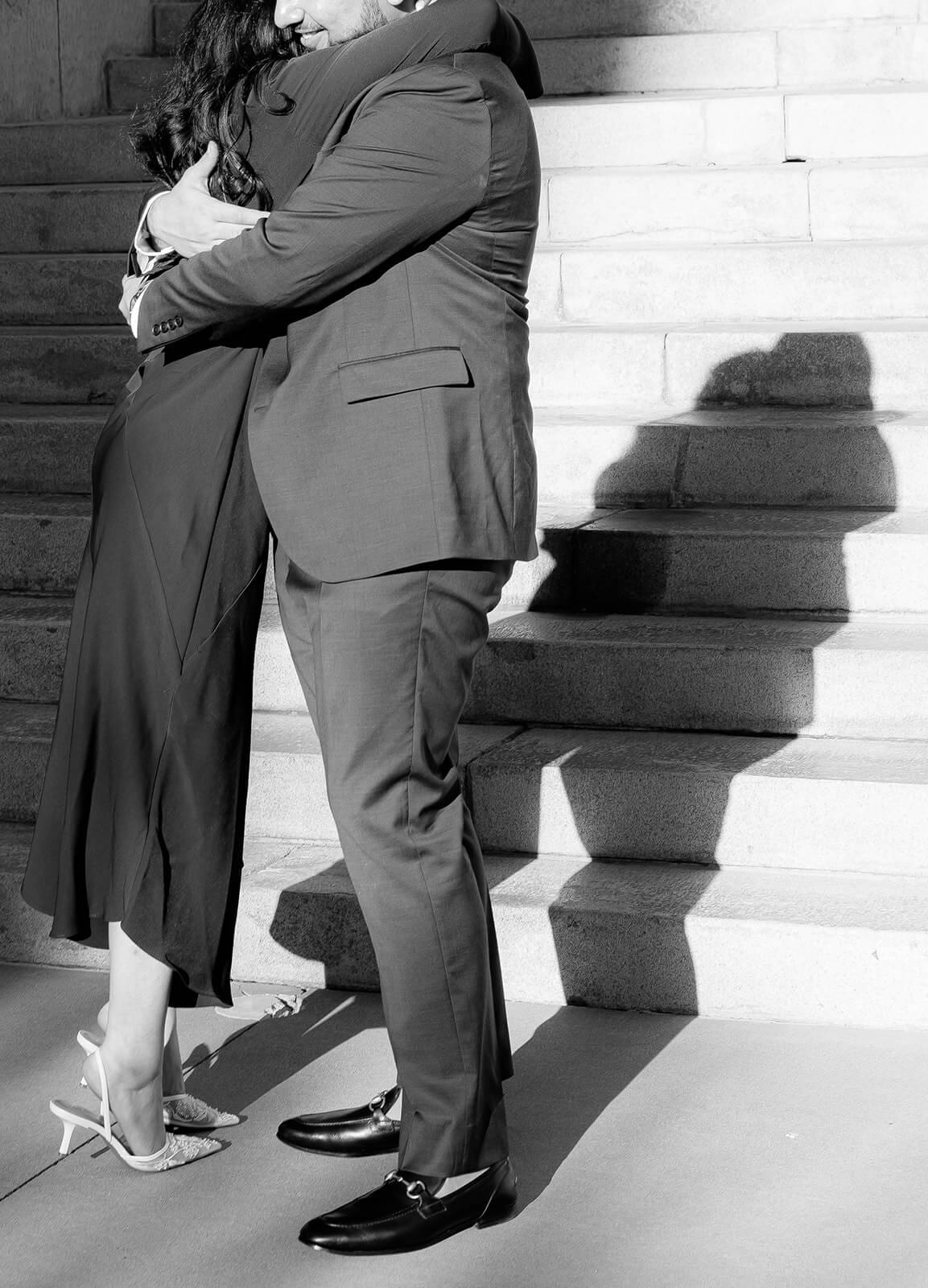 Black and white close-up of a couple standing together on stone steps, framed from the waist down, their hands intertwined.
