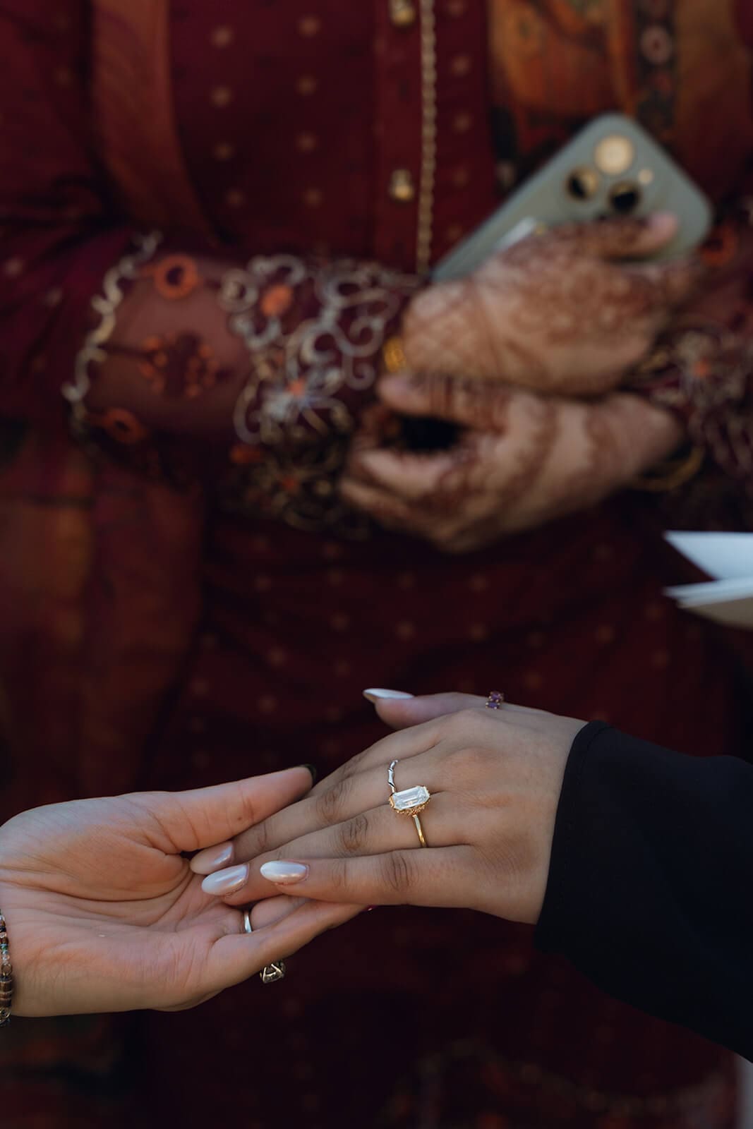 Close-up of a woman's hand adorned with henna and an engagement ring being held by another person's hand, with a phone visible in the background.
