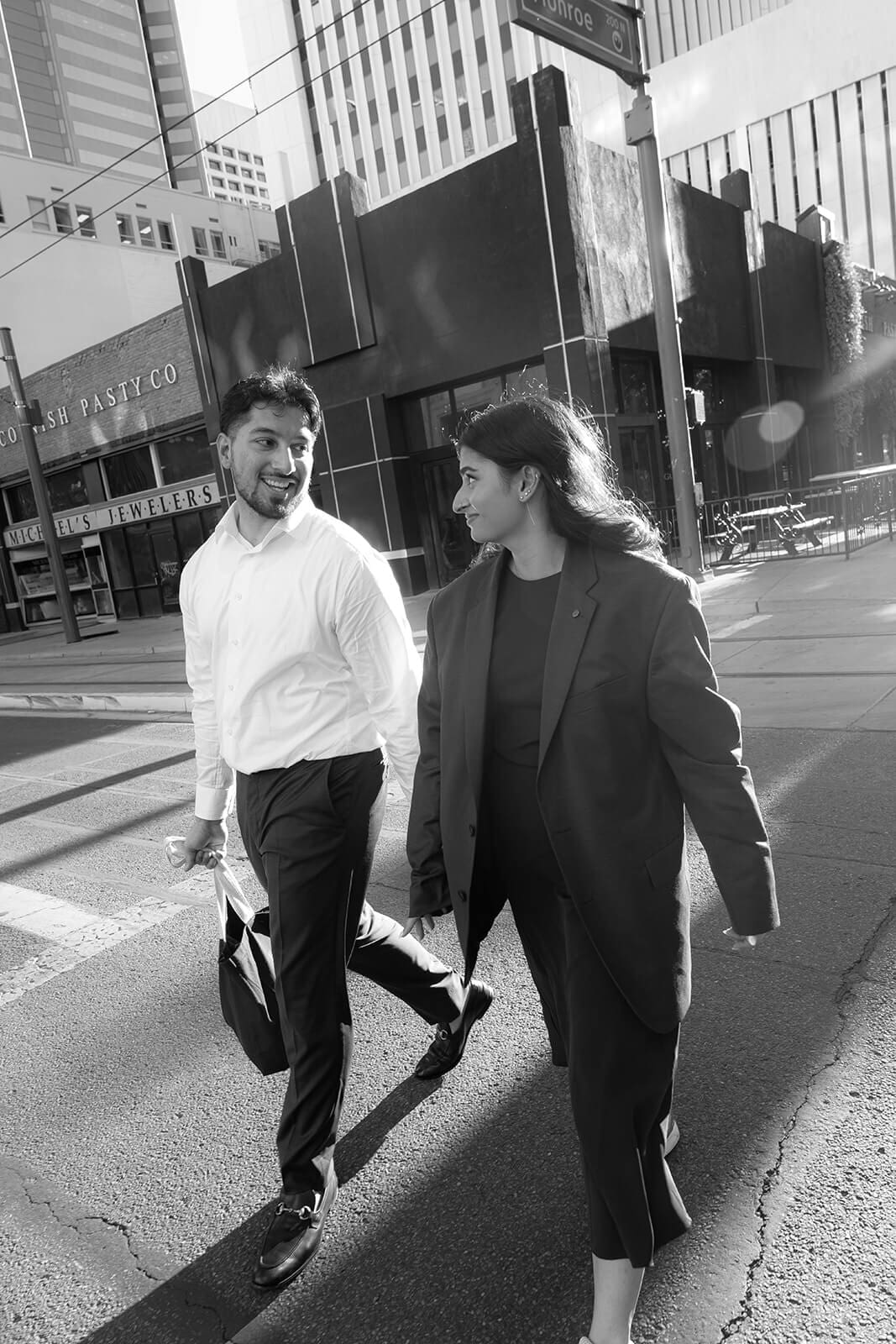 Black and white photo of the newly engaged couple walking confidently side by side down a city street, both dressed in stylish suits.
