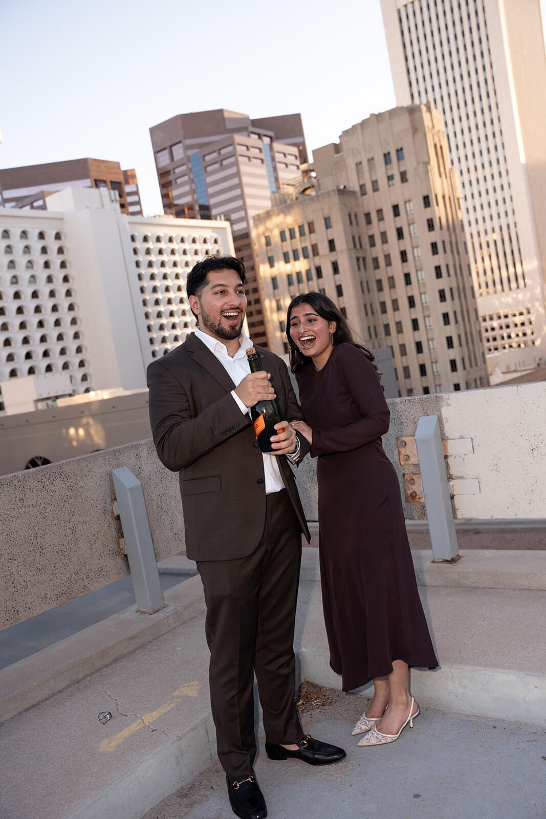 A couple celebrates their engagement on a rooftop with a city skyline behind them, the man popping a champagne bottle as the woman reacts with delight.
