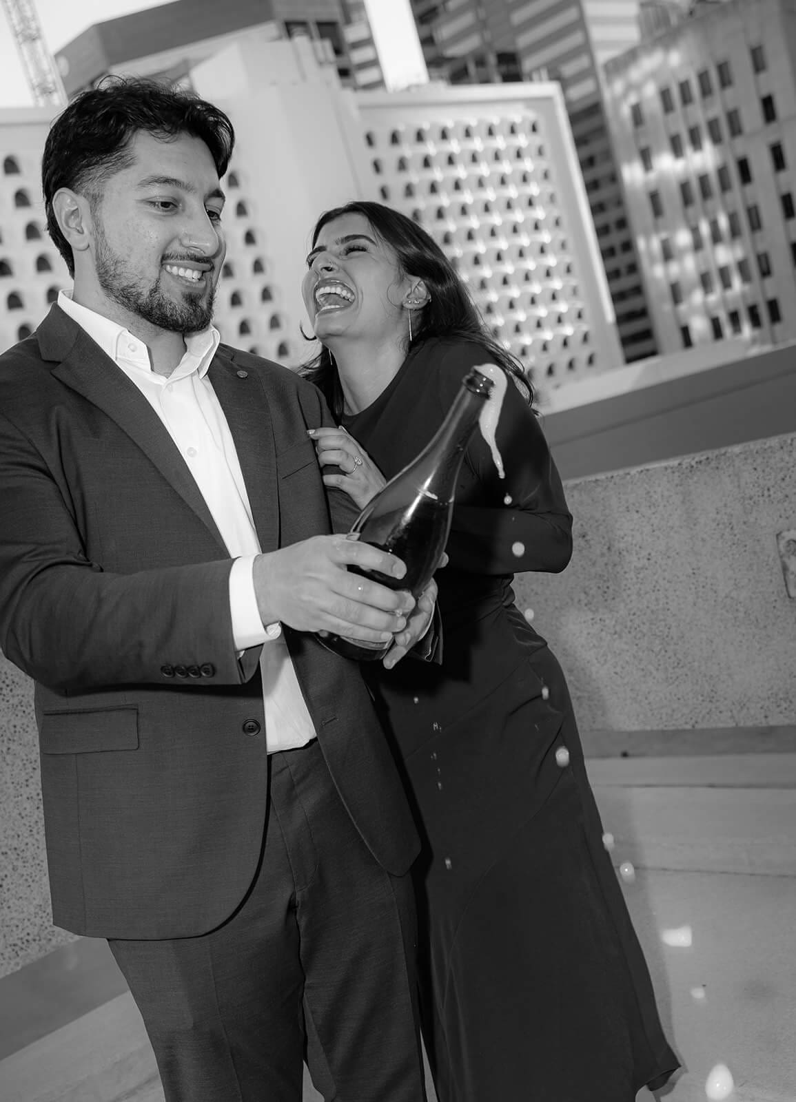 Black and white photo of a couple laughing together outdoors with a city skyline behind them, the man holding a champagne bottle as they celebrate their engagement.
