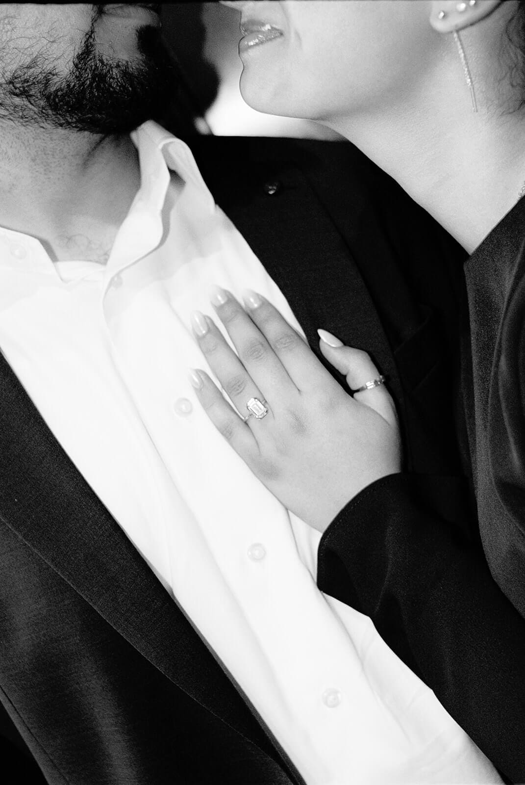 Black and white close-up of a woman's hand with a diamond engagement ring resting on a man's chest as they lean in close together, their faces partially visible.