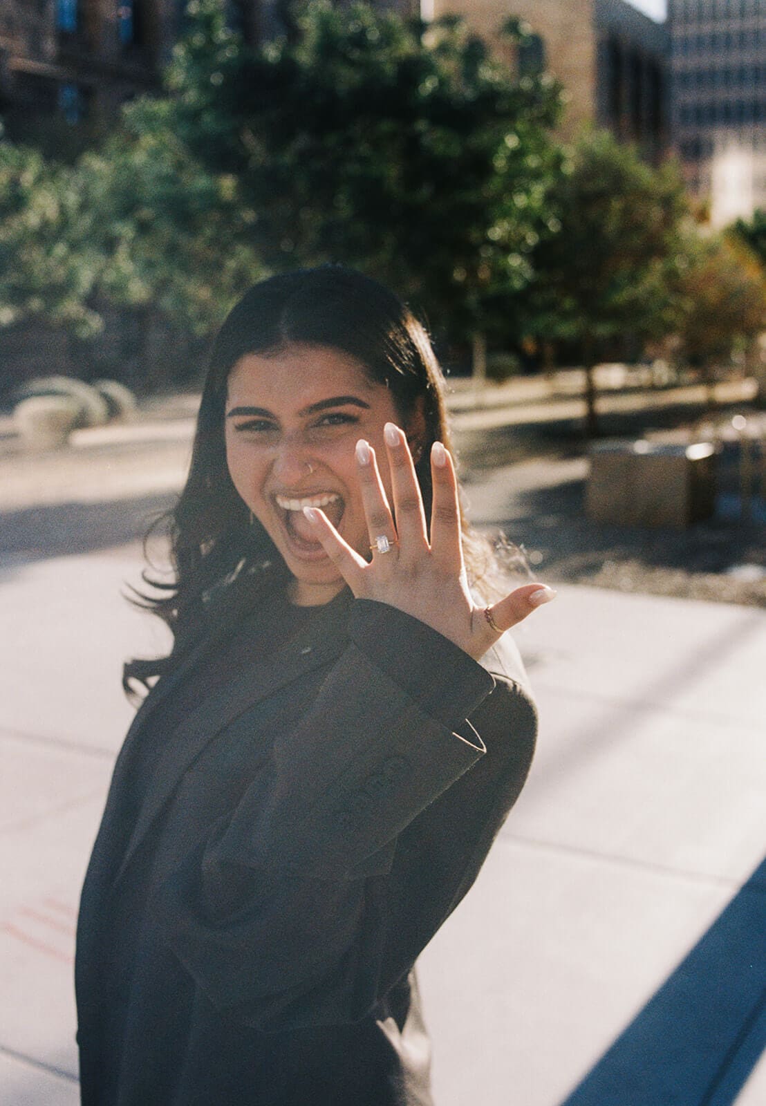 A woman grins widely at the camera while holding up her hand to show off her diamond engagement ring, standing outdoors in an urban setting.
