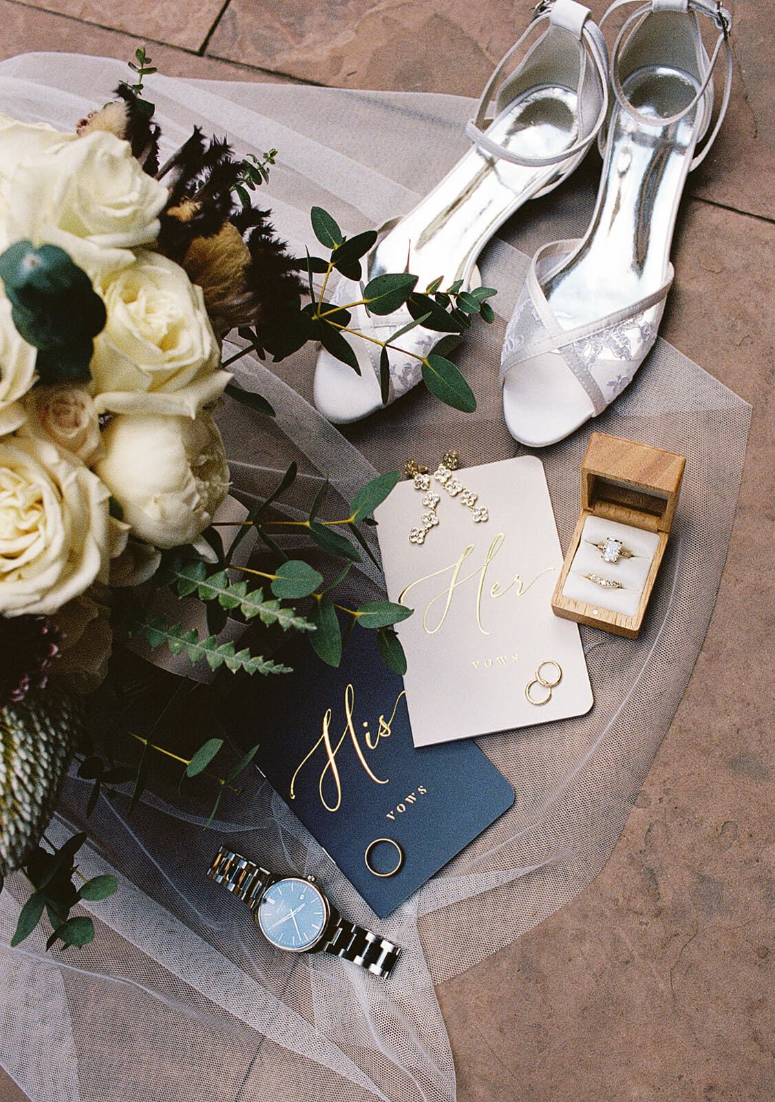 Flat lay of Sedona elopement details including white shoes, vow books, rings, jewelry, perfume, and a bridal bouquet on the red rocks.