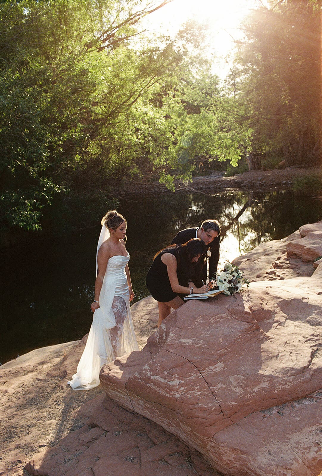 Bride and groom signing their marriage license with a witness during their Sedona elopement ceremony, surrounded by red rocks, greenery, and soft desert light.