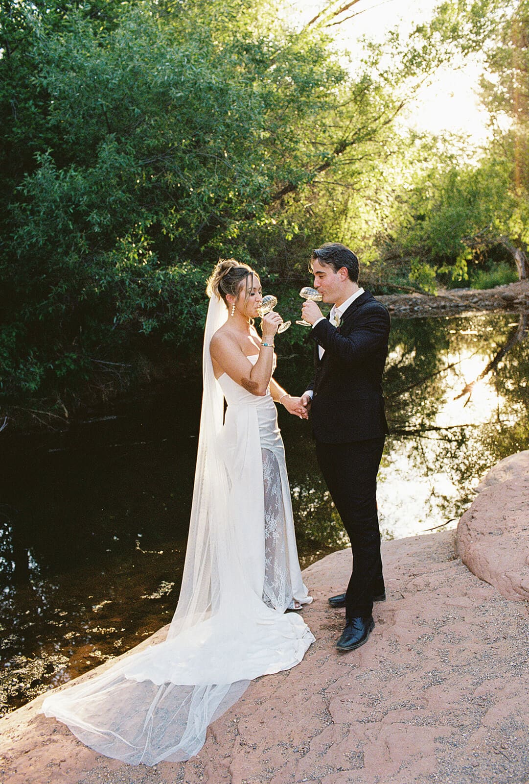 Bride and groom drinking champagne beside the creek after their Sedona elopement ceremony, with her lace train spread across the red rocks.