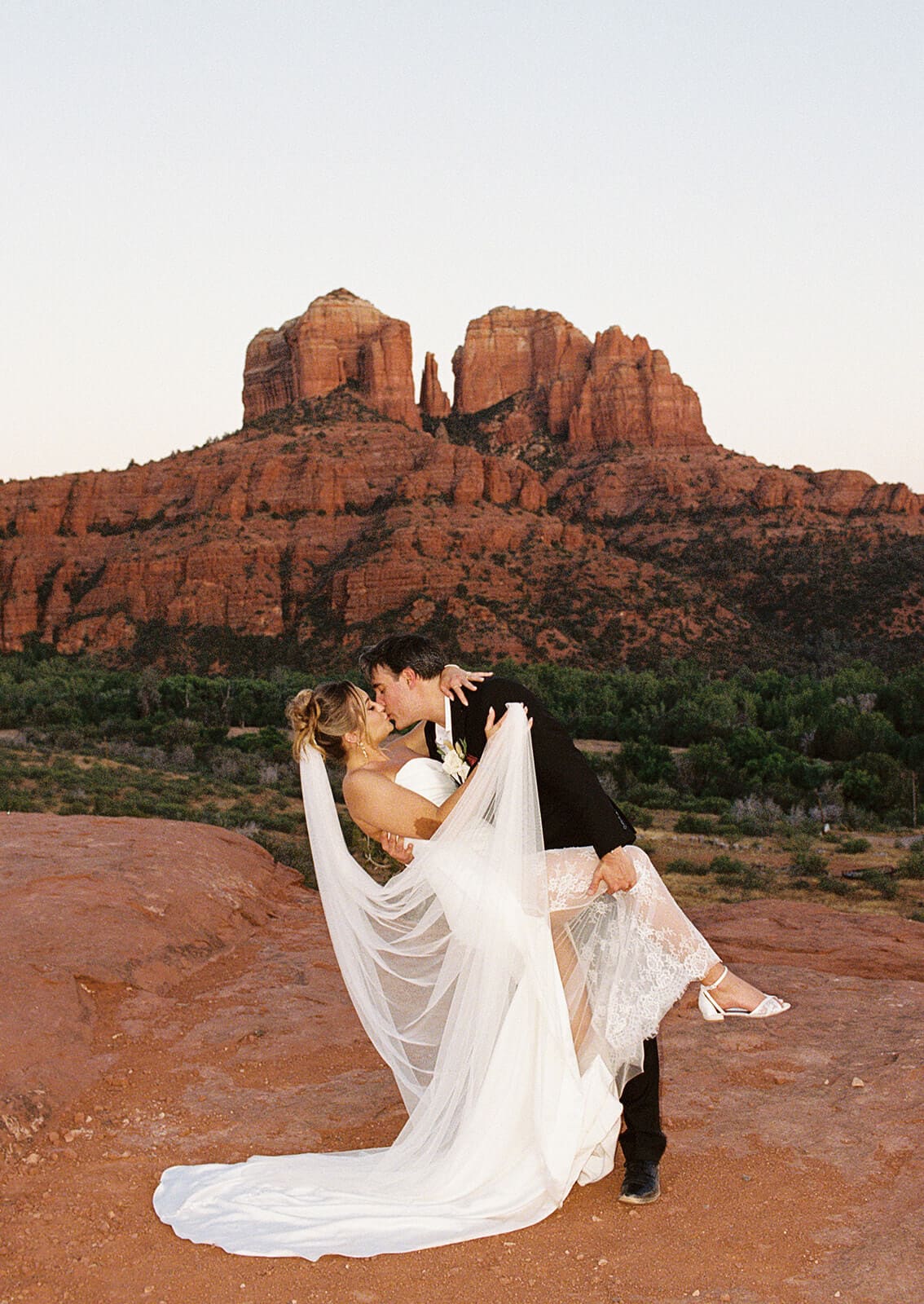 Bride and groom kissing on the red rocks during their Sedona elopement, with Cathedral Rock glowing in the background and her dress spread across the desert landscape.