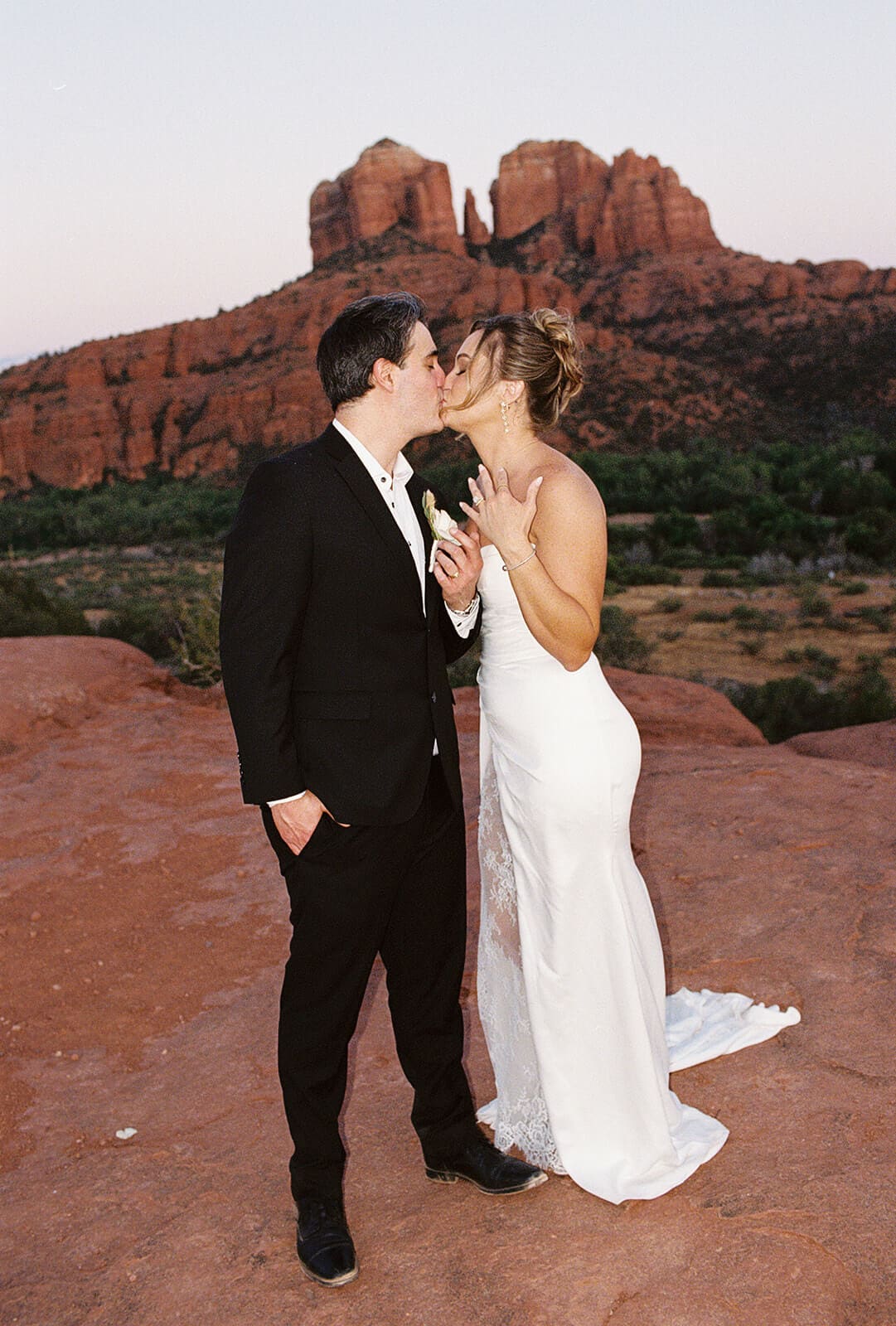 Bride and groom kissing on the red rocks after their Sedona elopement, with red mountains, desert greenery, and soft evening light behind them.