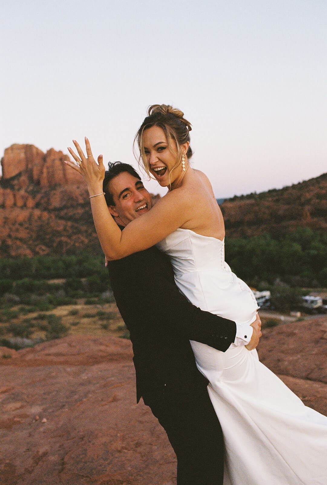 Groom lifting the bride in his arms on the red rocks during playful Sedona elopement portraits, with desert views and soft evening light behind them.