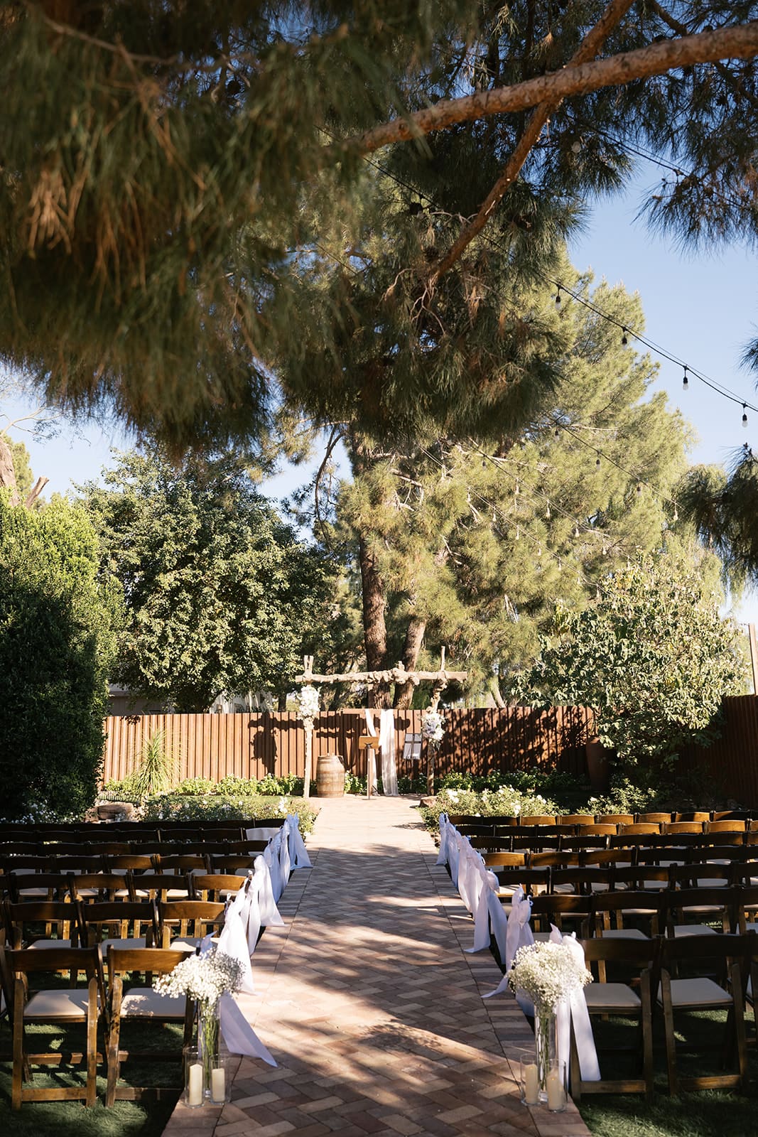Tree-lined ceremony aisle with wooden chairs and white fabric leading to the altar at Stonebridge Manor garden wedding venue in Mesa Arizona
