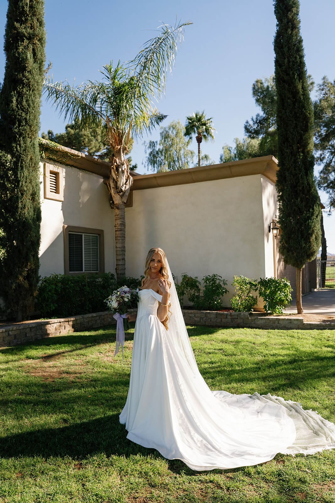 Bride standing in front of a stucco building surrounded by tall cypress trees at Knotty Barn wedding venue in Queen Creek Arizona