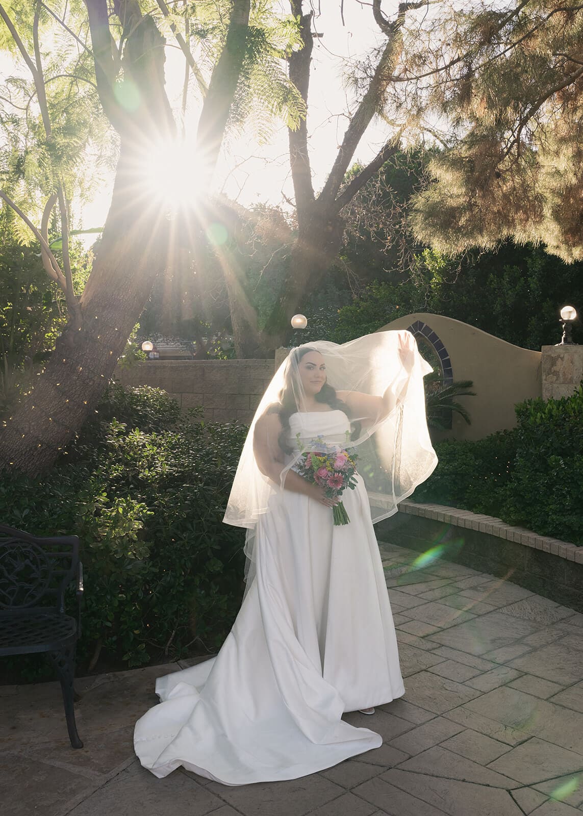 Bride standing alone in golden backlit sunlight with her veil lifting in the breeze at The Wright House garden wedding venue in Mesa Arizona
